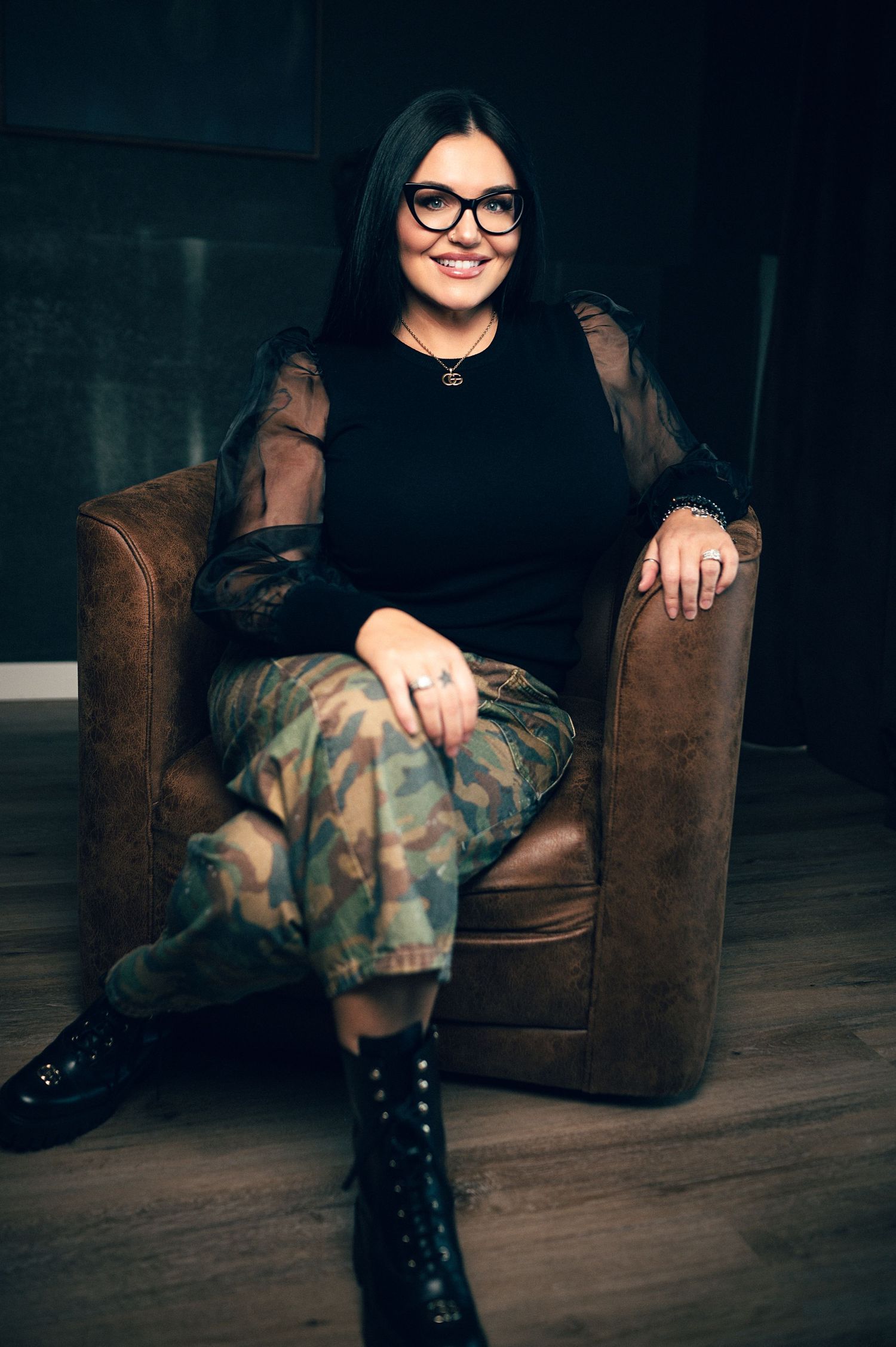 Smiling woman with glasses in black top and camo pants seated in brown leather chair.