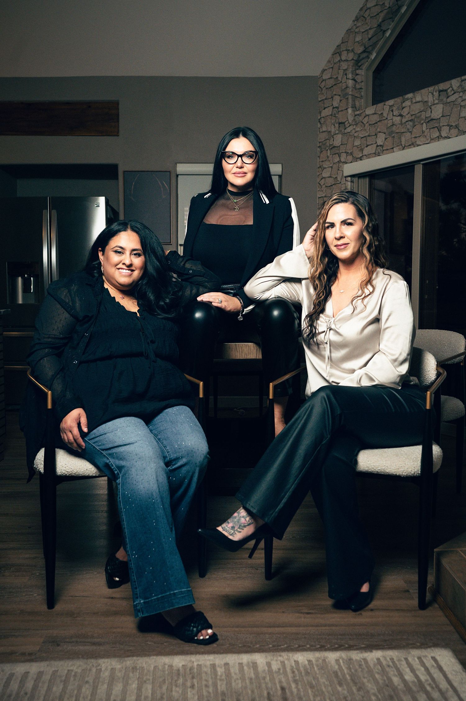 Three women posing elegantly in a modern interior, two seated in chairs and one standing, dressed in stylish outfits.