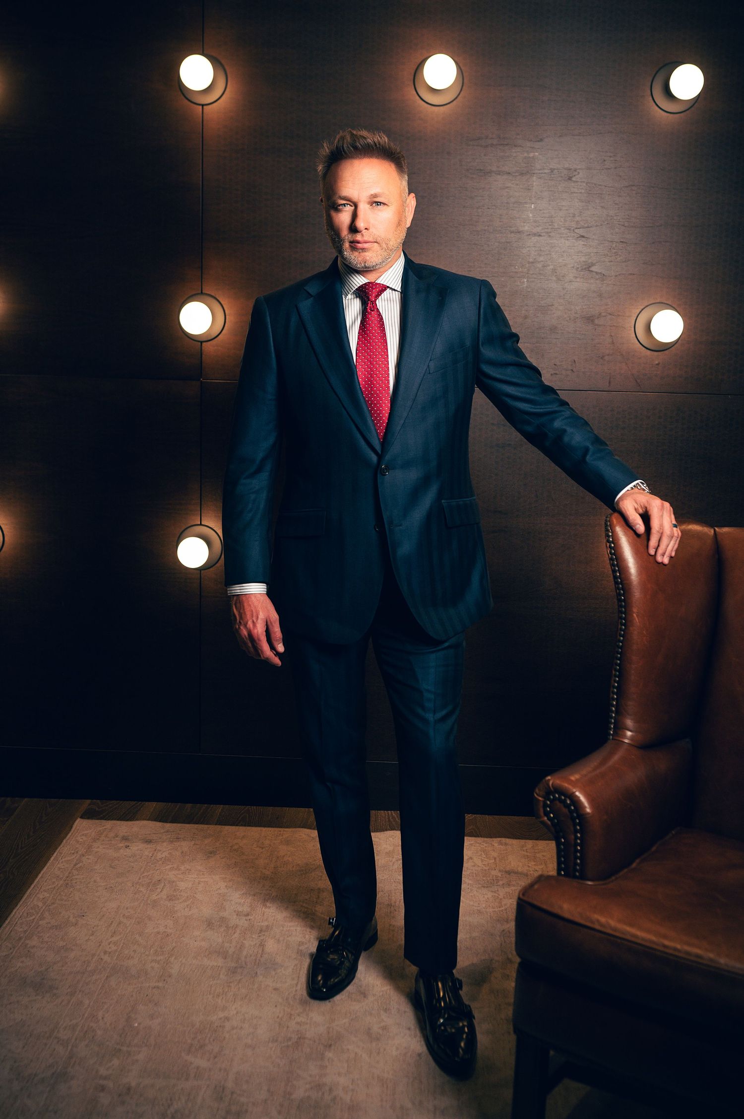 Professional man in navy suit and red tie standing confidently beside leather chair against dark wall with spotlights.