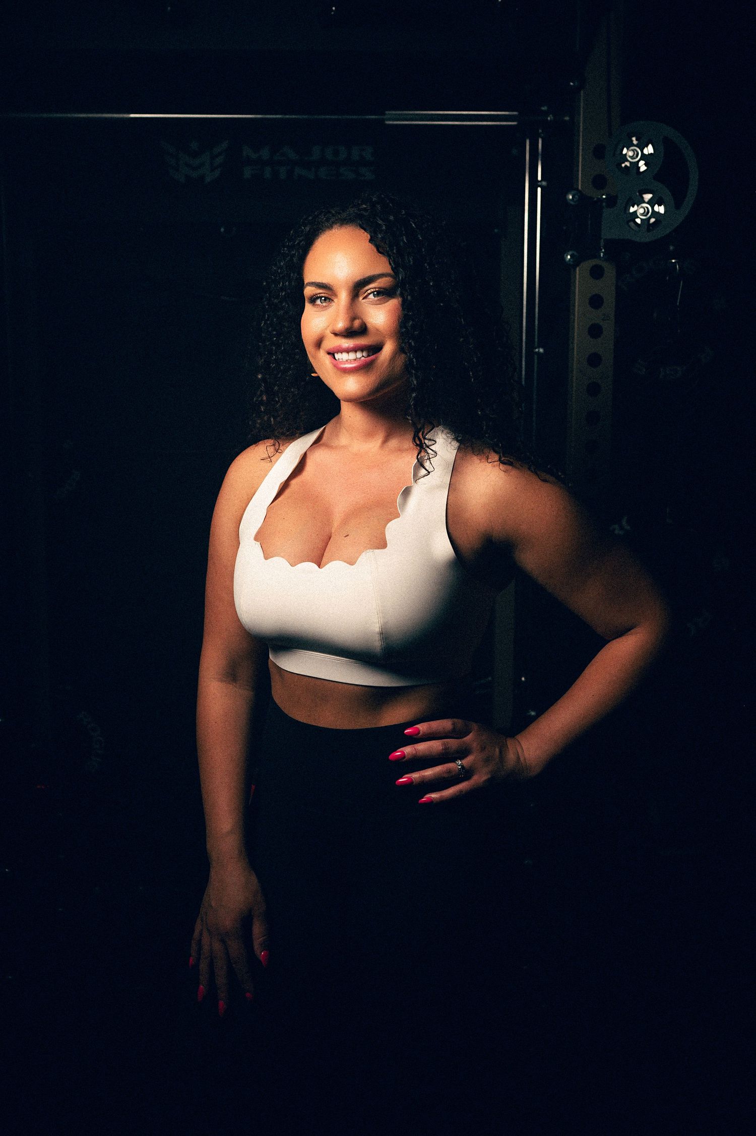Smiling woman in white sports bra poses confidently in a dark gym setting with curly hair and pink nails.