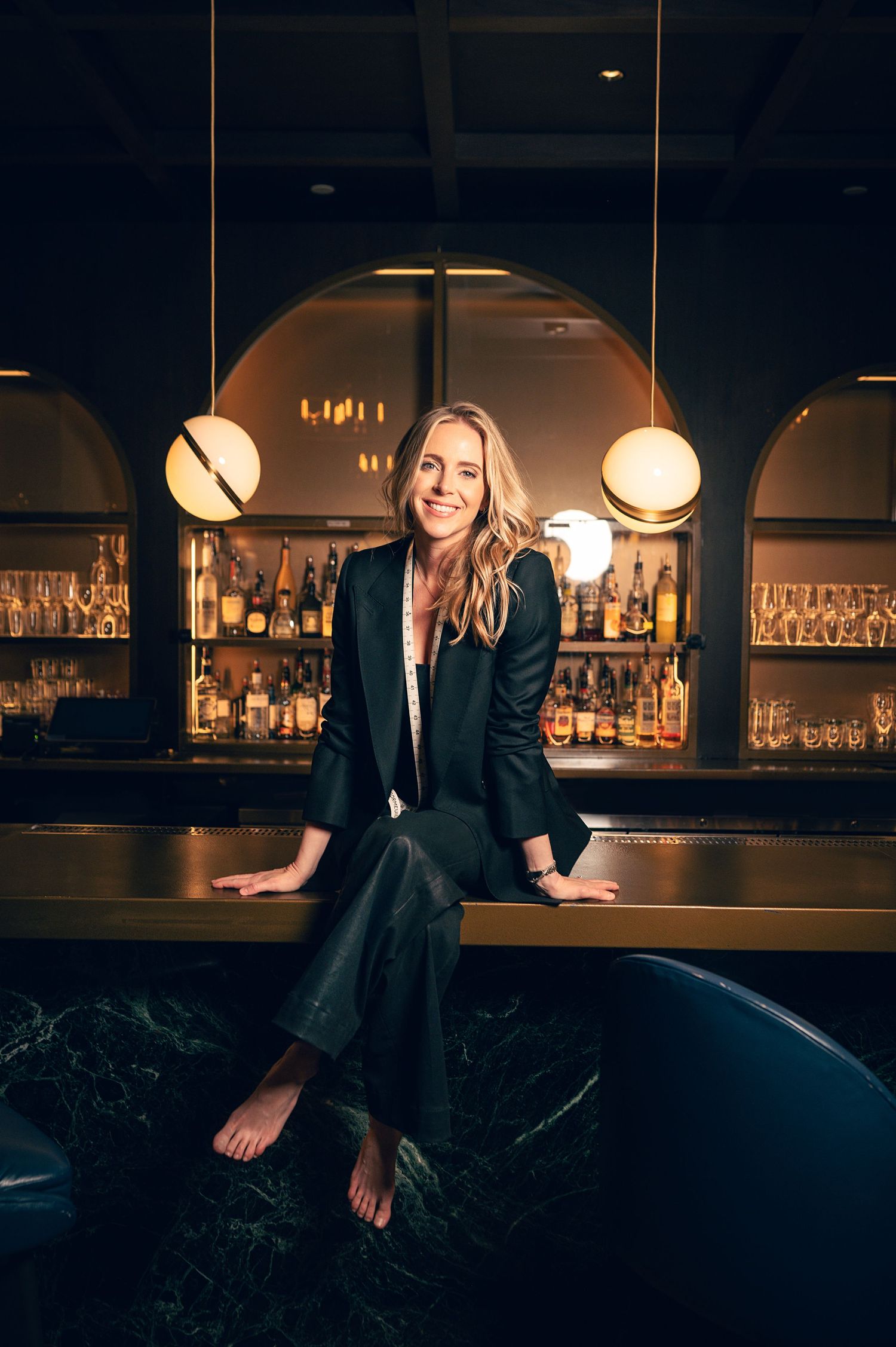 Smiling blonde woman in black blazer sitting barefoot on bar counter in elegant dimly lit cocktail lounge.