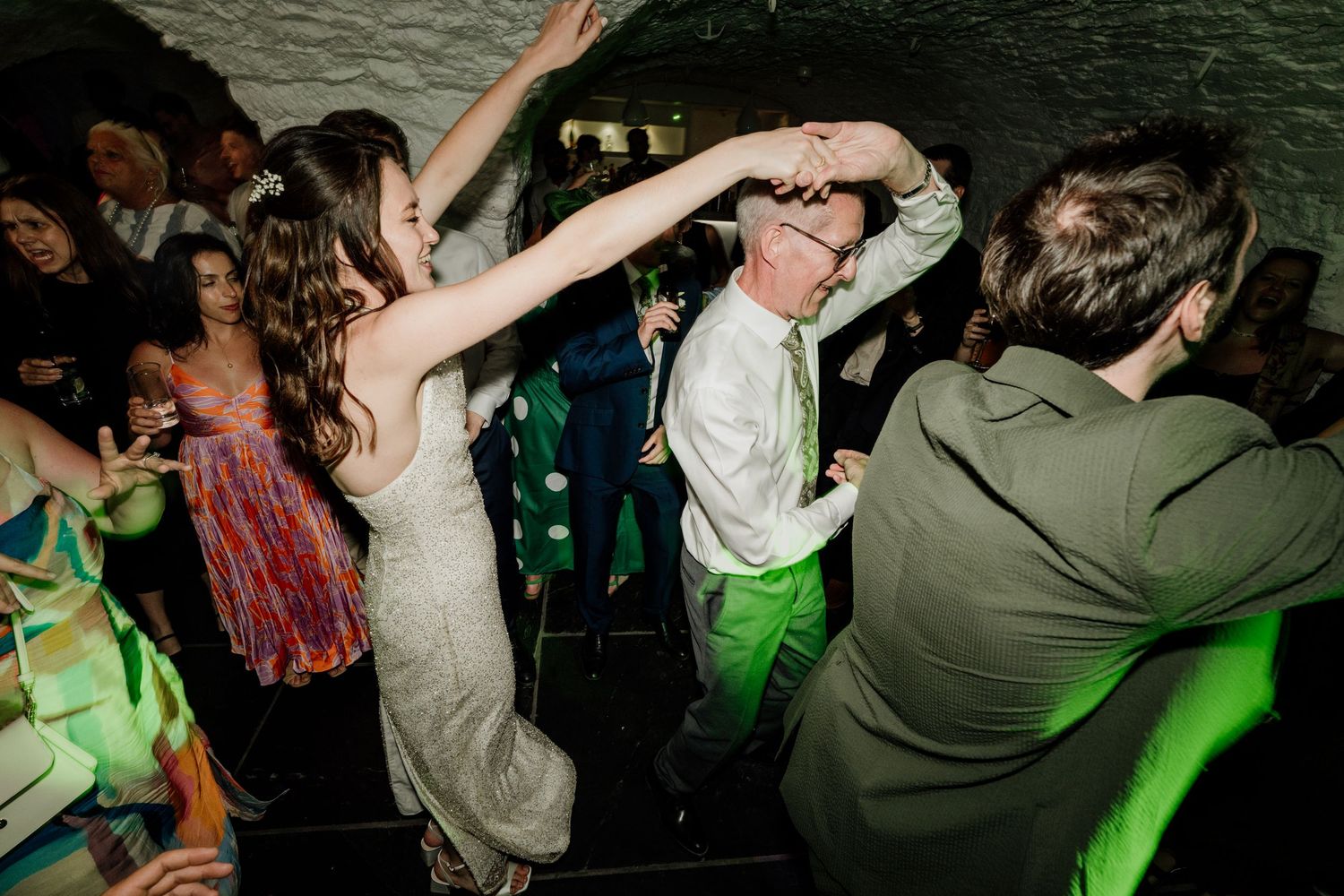 Bride and groom dancing energetically at a lively wedding reception with green lighting and guests in background.