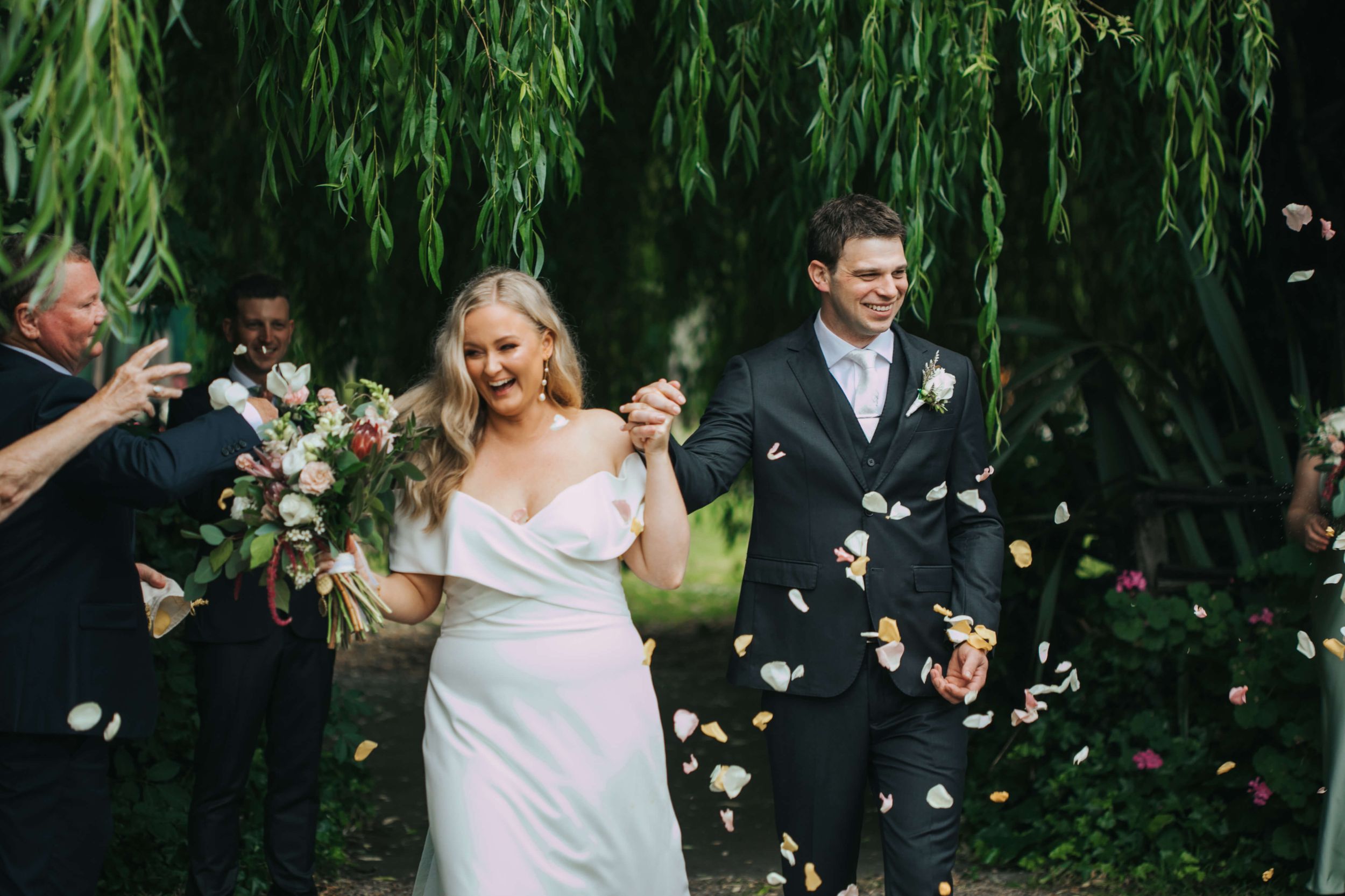 Joyful newlyweds walking hand in hand through a confetti shower under lush green weeping willow trees.