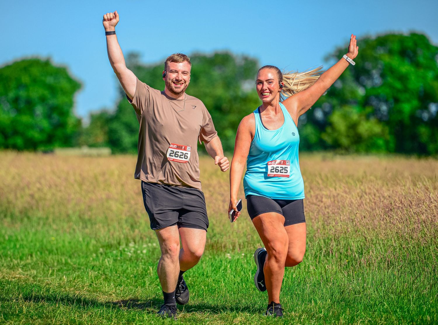 Smiling man and woman with race bibs celebrate while running through sunny green field together.
