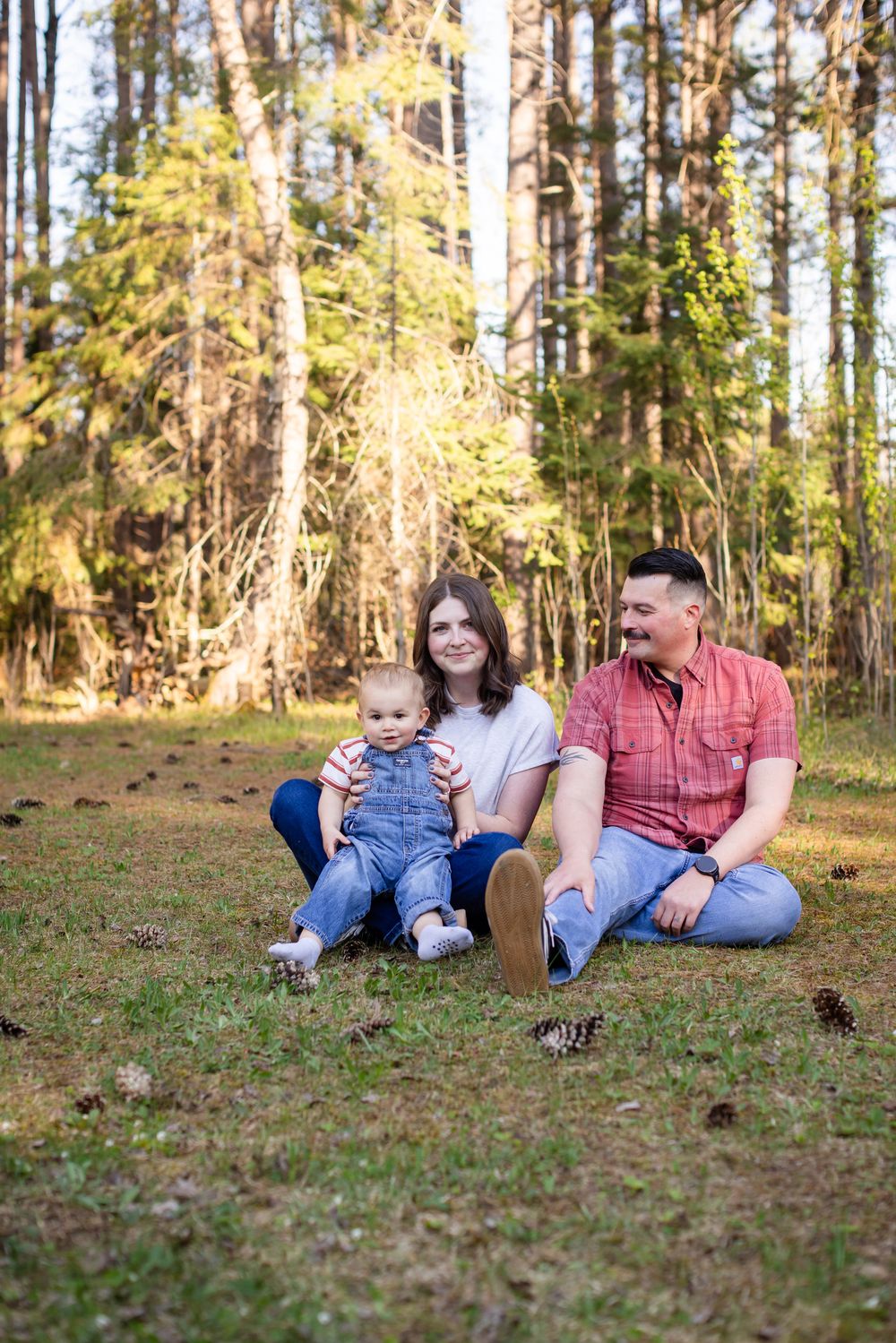 Young family with baby sitting on grass in sunny pine forest.