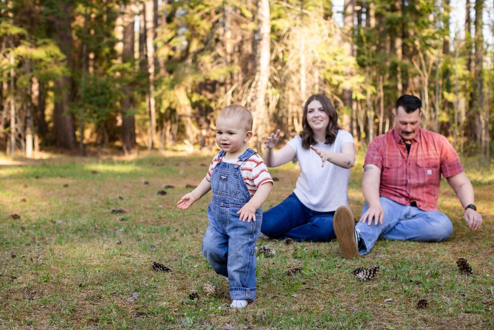 A happy young family with a toddler playing outdoors in a sunny pine forest, parents sitting on grass smiling.