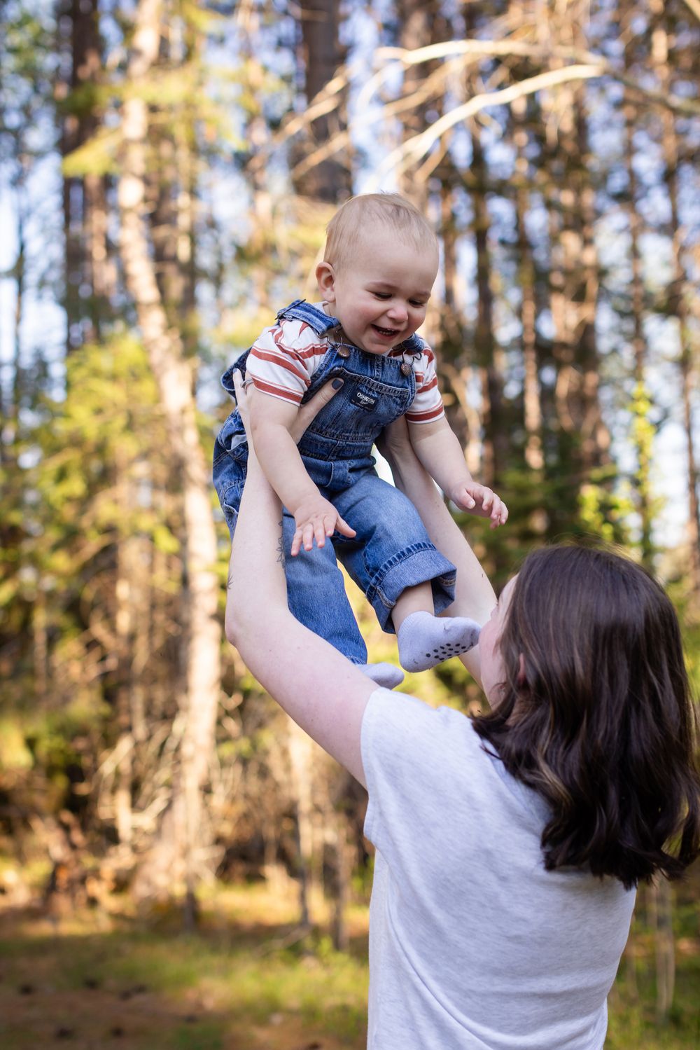 A joyful mother lifts her laughing baby in denim overalls high in the air outdoors in a sunlit forest.