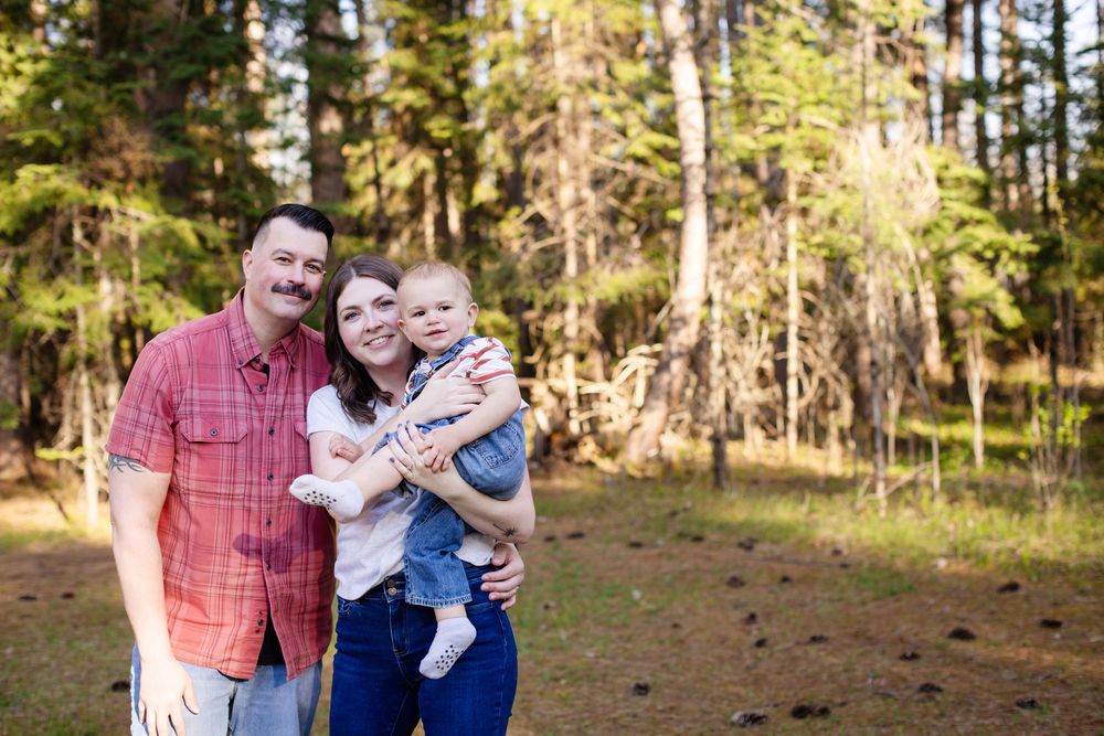 A happy family of three poses outdoors in a sunlit forest, with parents holding their baby in a scenic woodland setting.