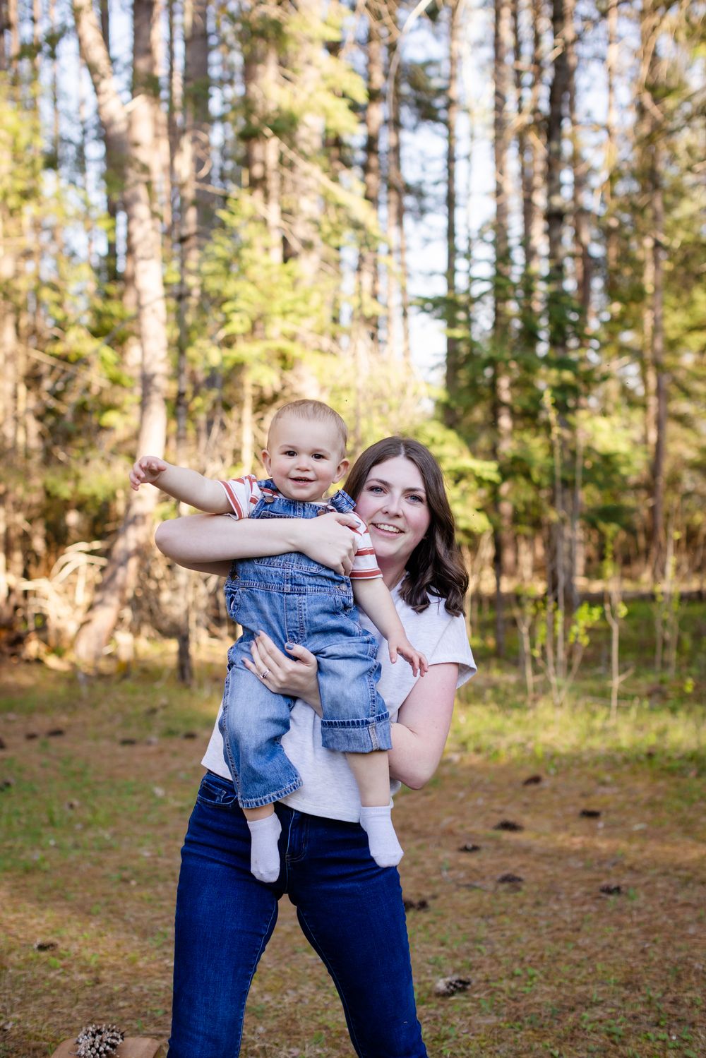 Smiling mother holding happy baby outdoors in sunny pine forest.