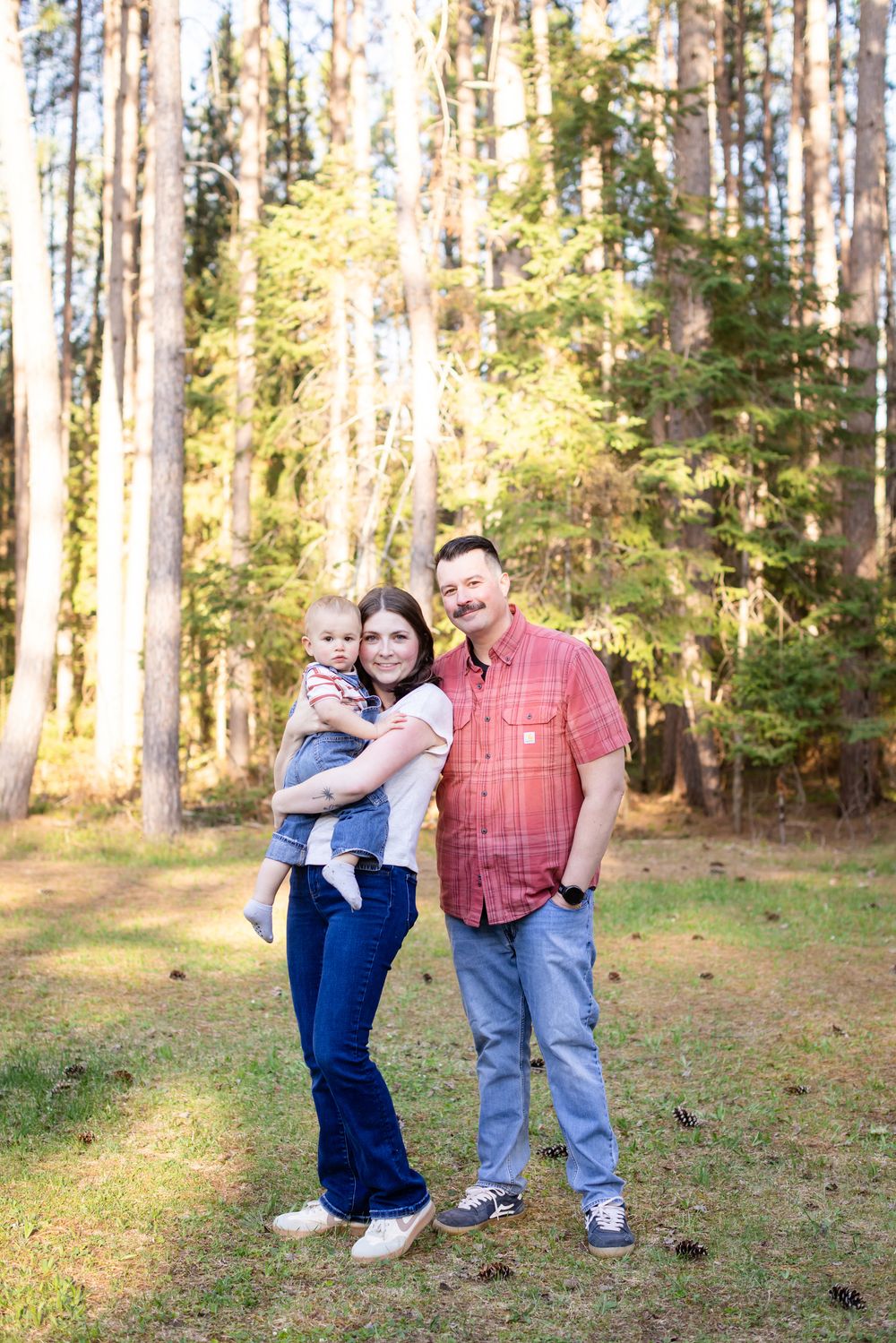 Family of three posing outdoors in a sunlit forest, parents smiling with baby in arms, wearing casual jeans and shirts.