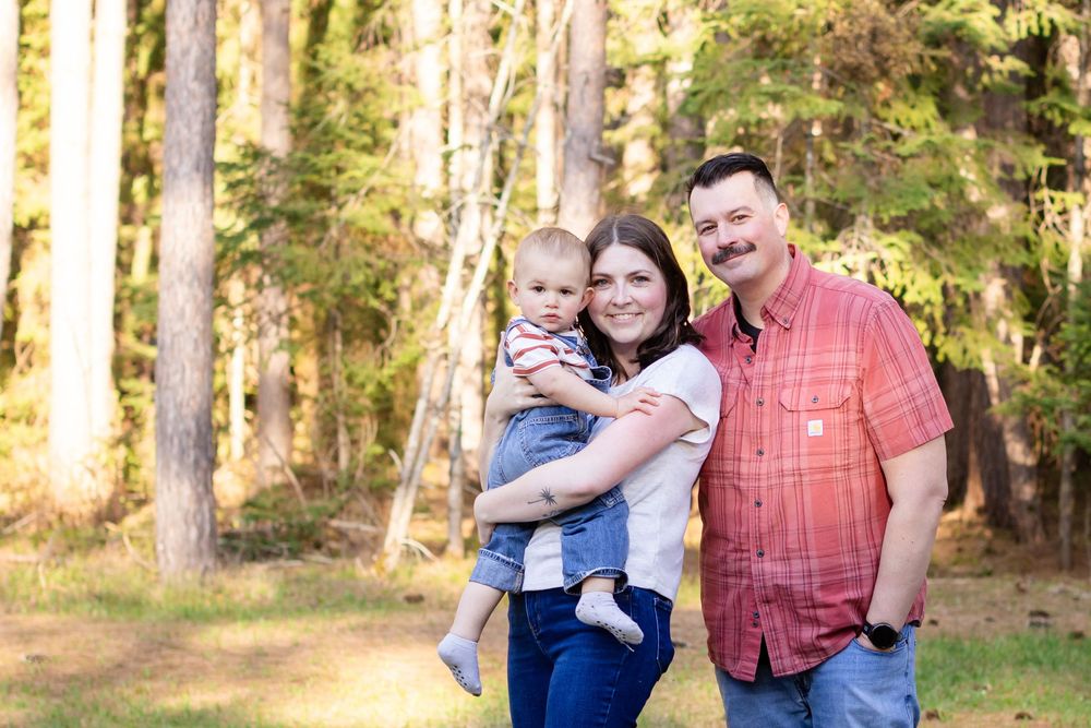A happy young family with a baby poses outdoors in a sunlit forest clearing for a professional portrait.