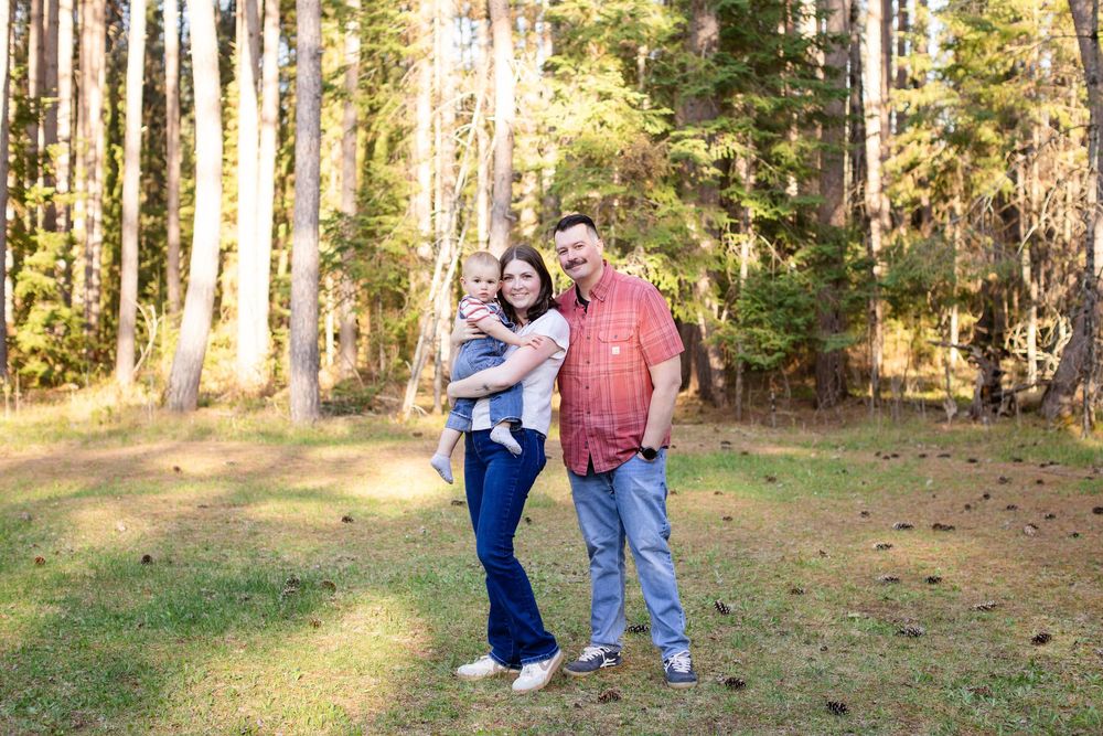 A happy family of three posing together in a sunlit forest clearing, with tall pine trees in the background.