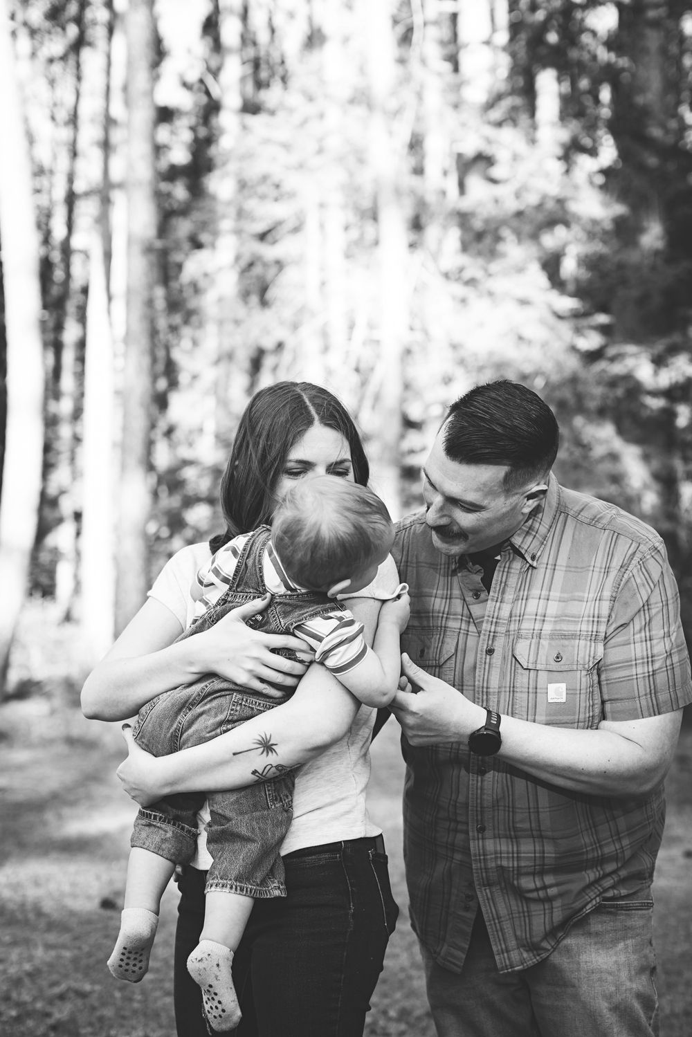 Black and white photo of a young family with mother holding baby and father smiling in a forest setting.
