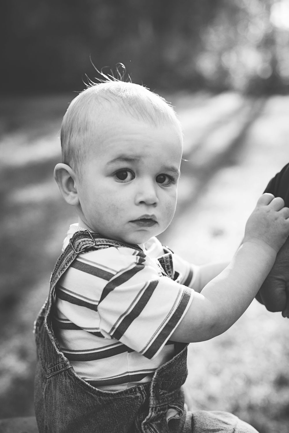 Black and white portrait of a curious toddler boy in striped shirt and overalls outdoors.