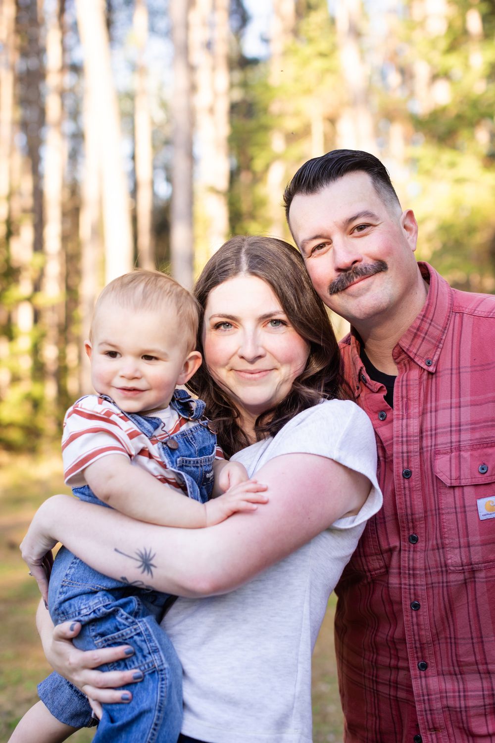 A happy young family with a toddler boy poses outdoors in a pine forest for a portrait photo.