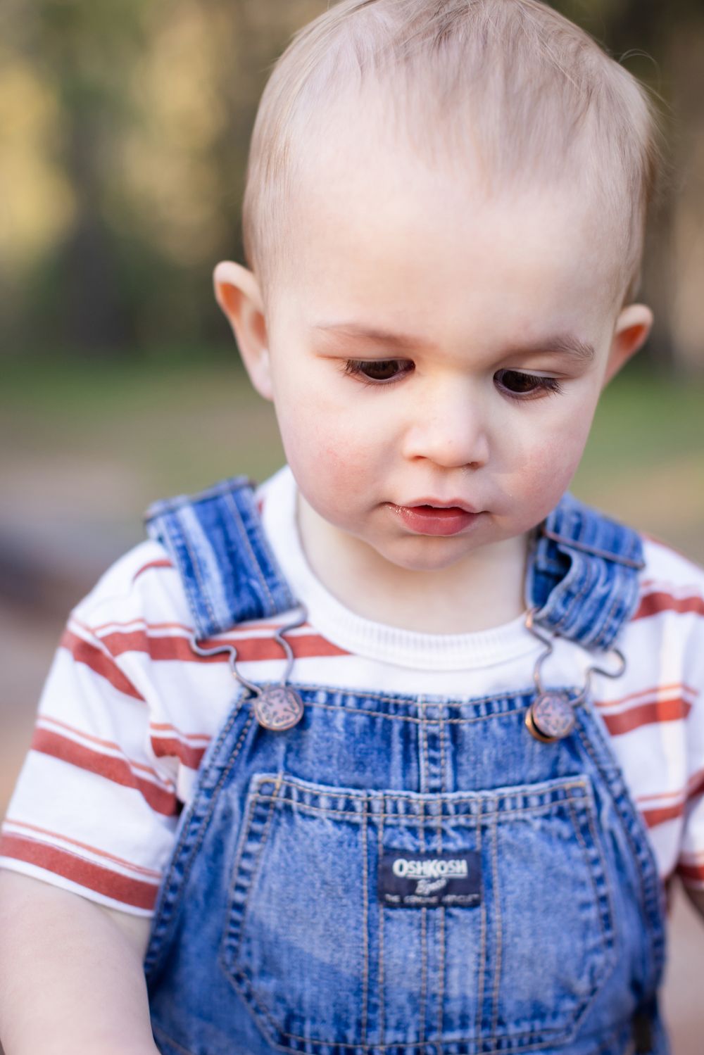 An adorable toddler in OshKosh denim overalls and striped shirt looking down outdoors.