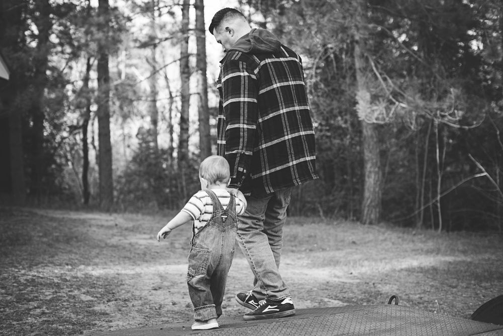Black and white photo of a father and toddler walking together on a path in a wooded outdoor setting.