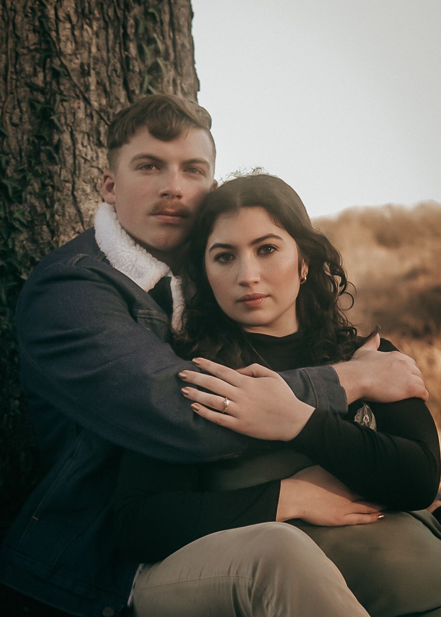 A young couple sitting against a tree, embracing tenderly in a natural outdoor setting.