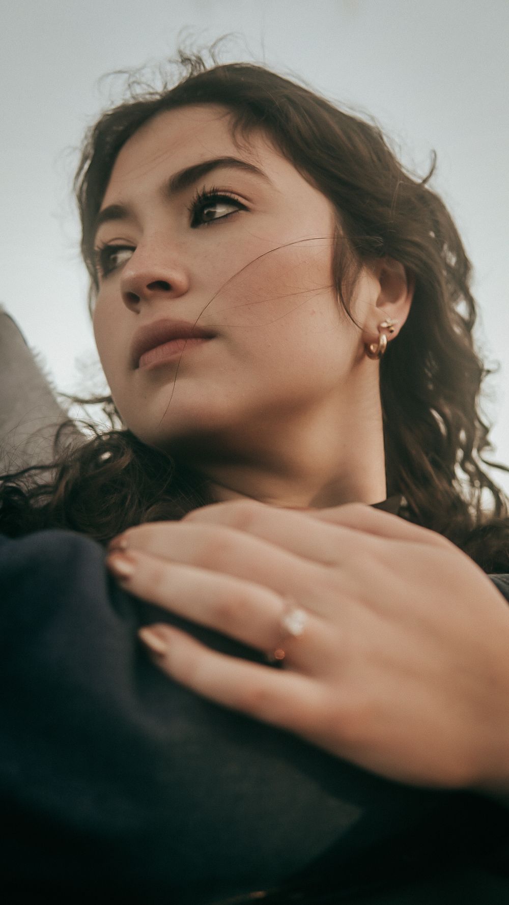 Young woman with curly hair and diamond ring gazes upward, wearing gold earrings in a moody portrait shot.