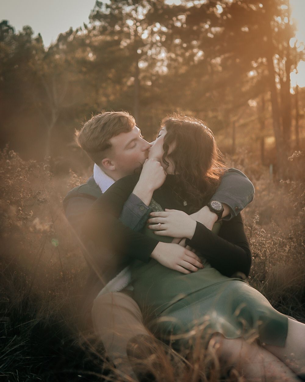 Couple sharing a romantic kiss at golden hour in a sunlit forest field, man cradling woman's face tenderly.