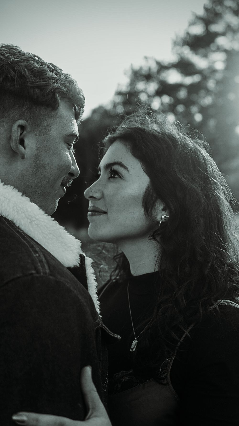 Black and white romantic couple gazing at each other outdoors, woman with curly hair wearing necklace, man in sherpa jacket.