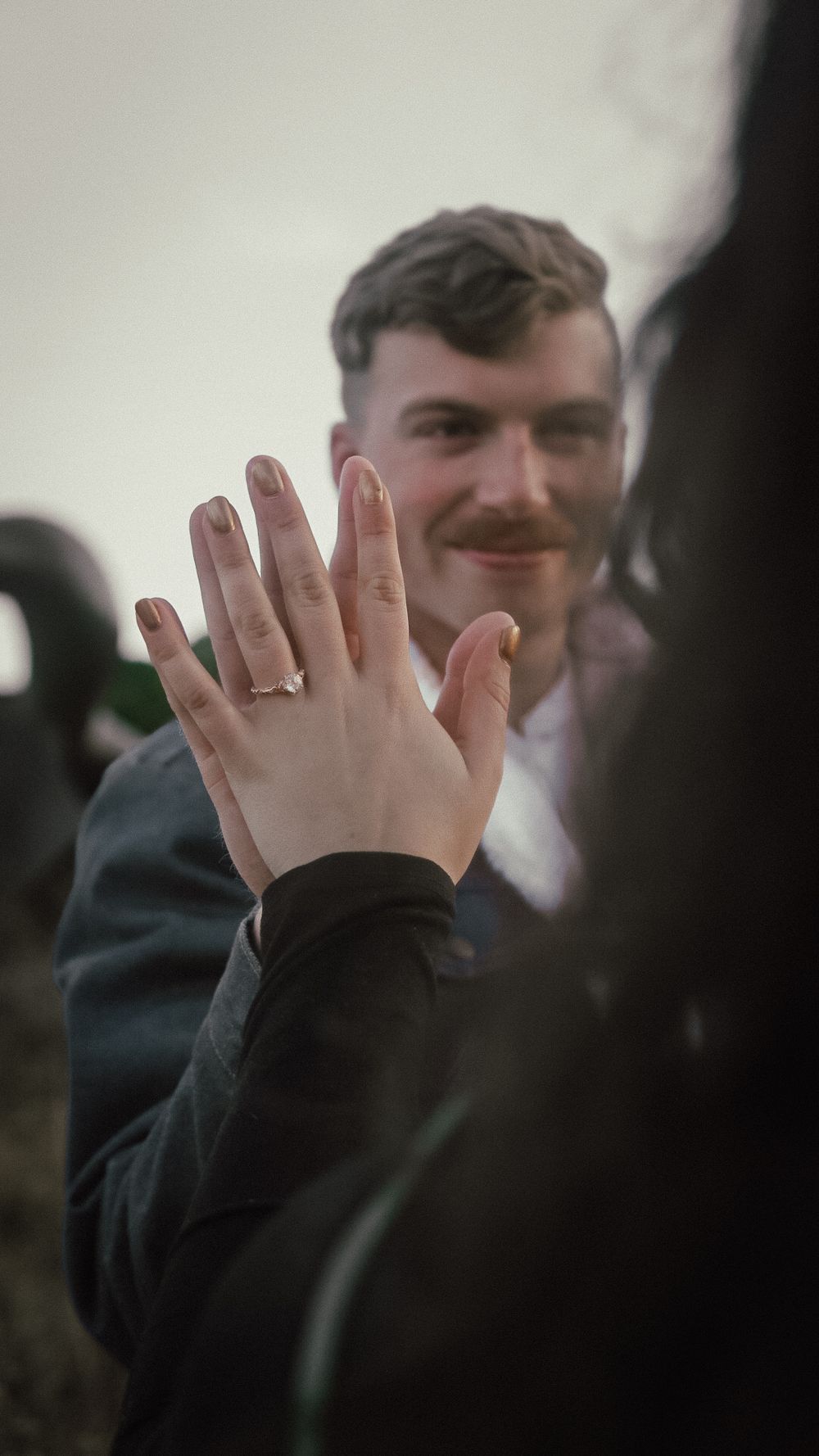 Smiling man with mustache behind a woman showing off her engagement ring outdoors.