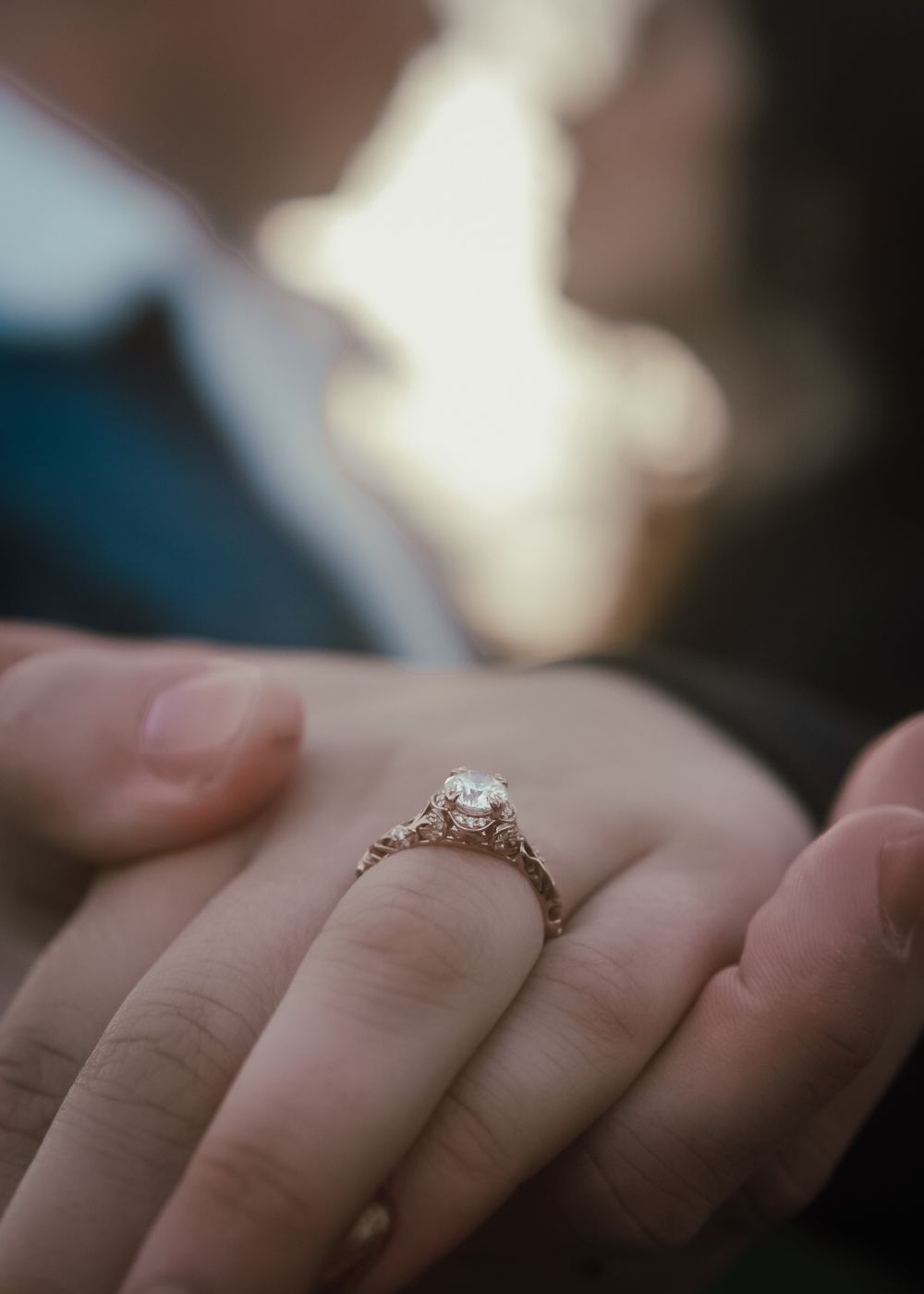 Close-up of an elegant rose gold diamond engagement ring on a womans finger with couple kissing.