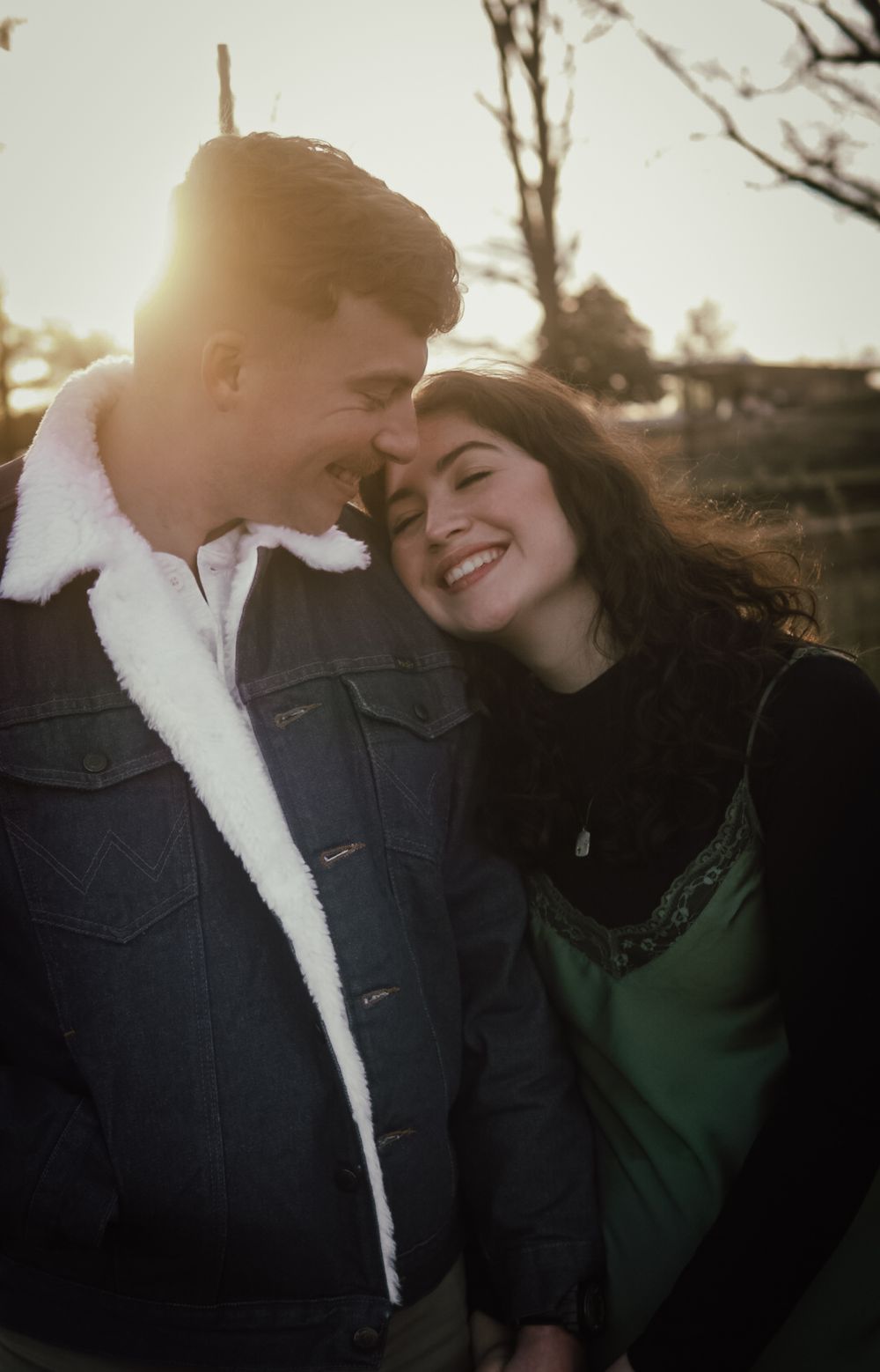 A couple smiles and leans together outdoors at golden hour, man in sherpa jacket, woman in green dress.