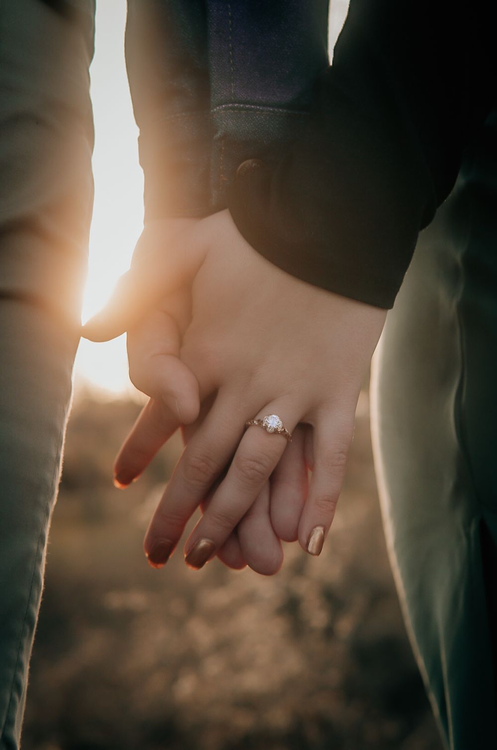 Couple holding hands at sunset, woman wearing a diamond engagement ring with red nail polish, romantic proposal moment.
