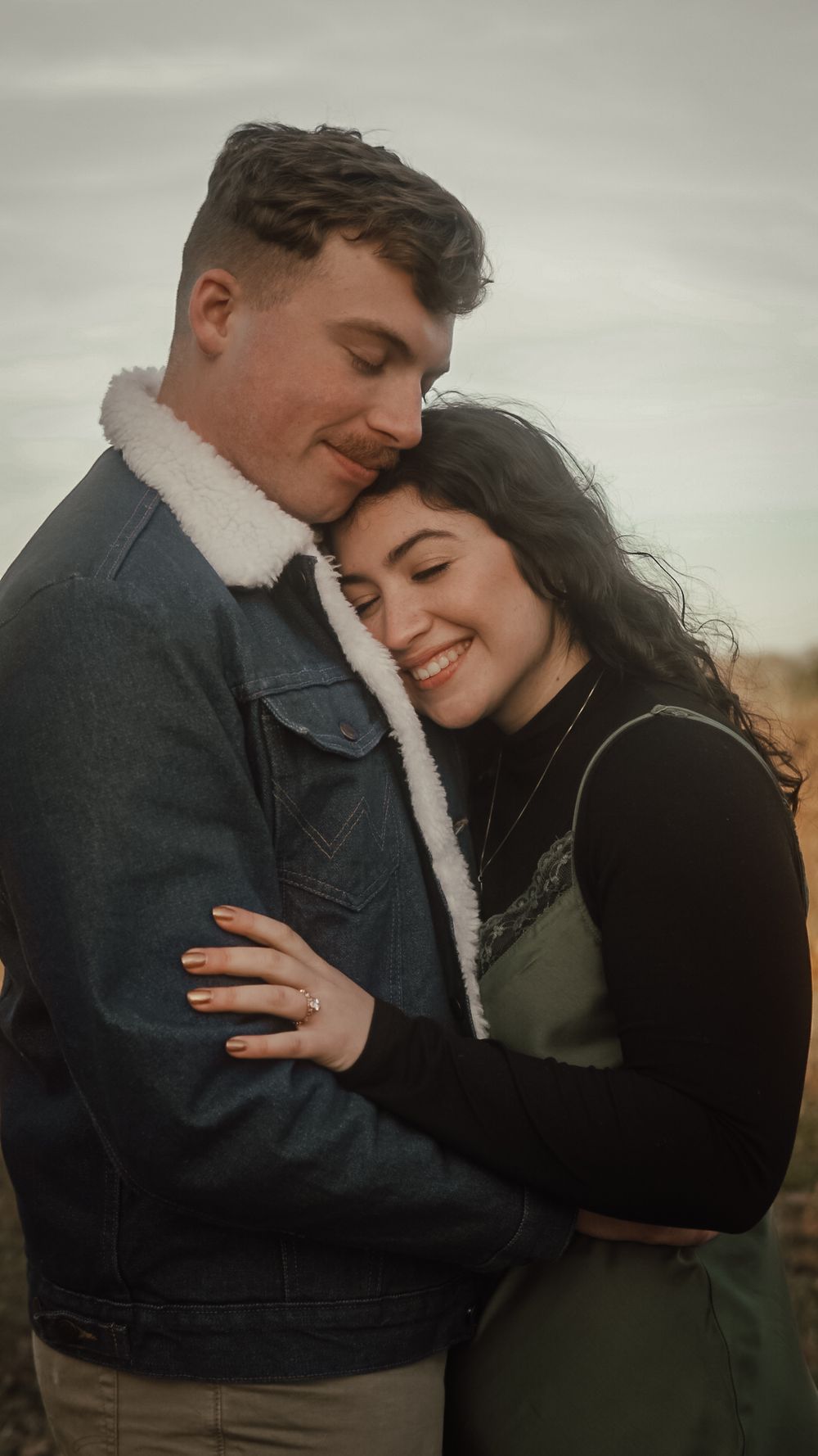 A smiling couple embraces outdoors, woman in green dress leans on man in denim sherpa jacket, sharing a tender romantic moment.