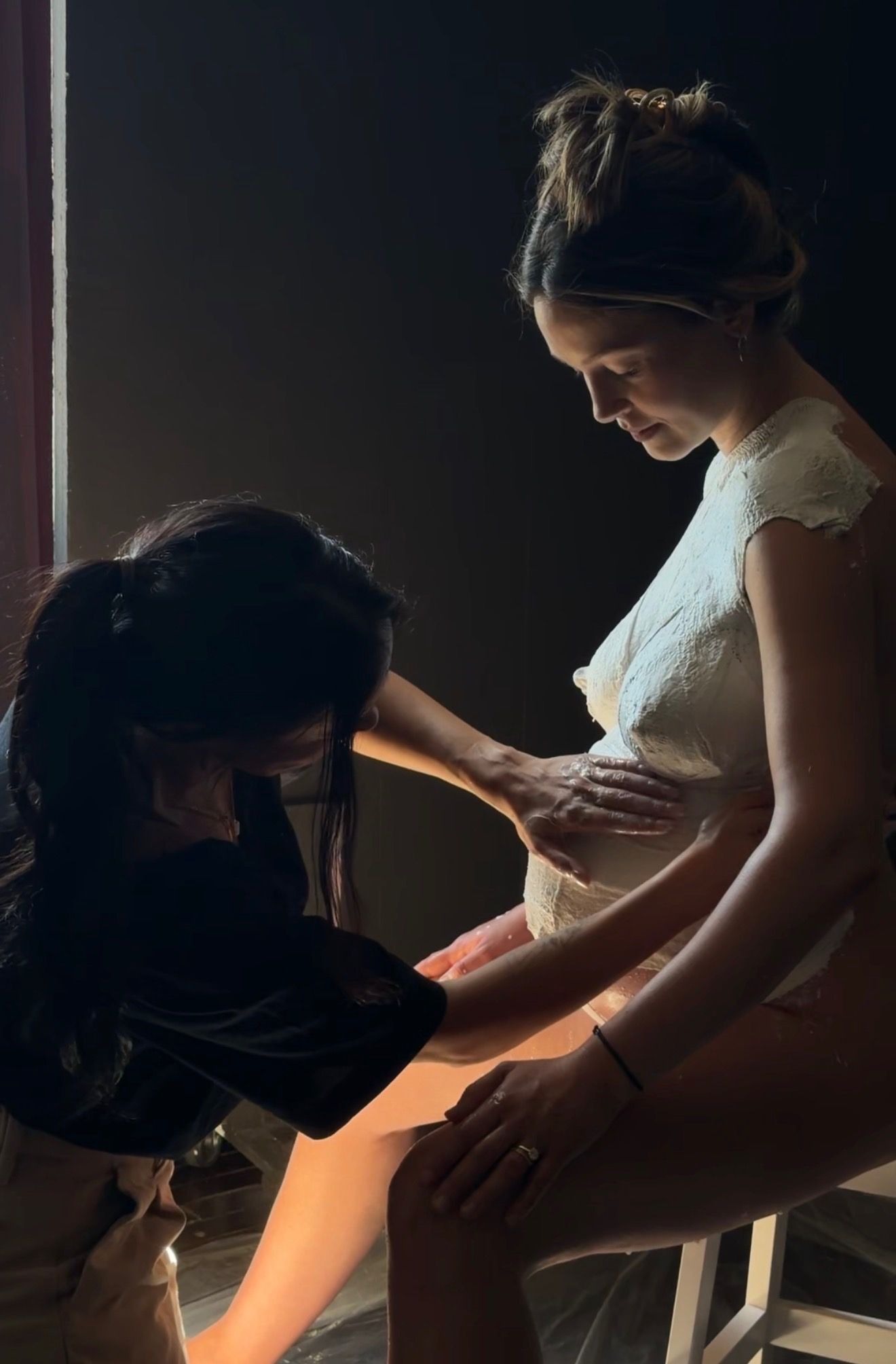 Two women in a dimly lit studio, one kneeling adjusting the other's white dress during a photo shoot.