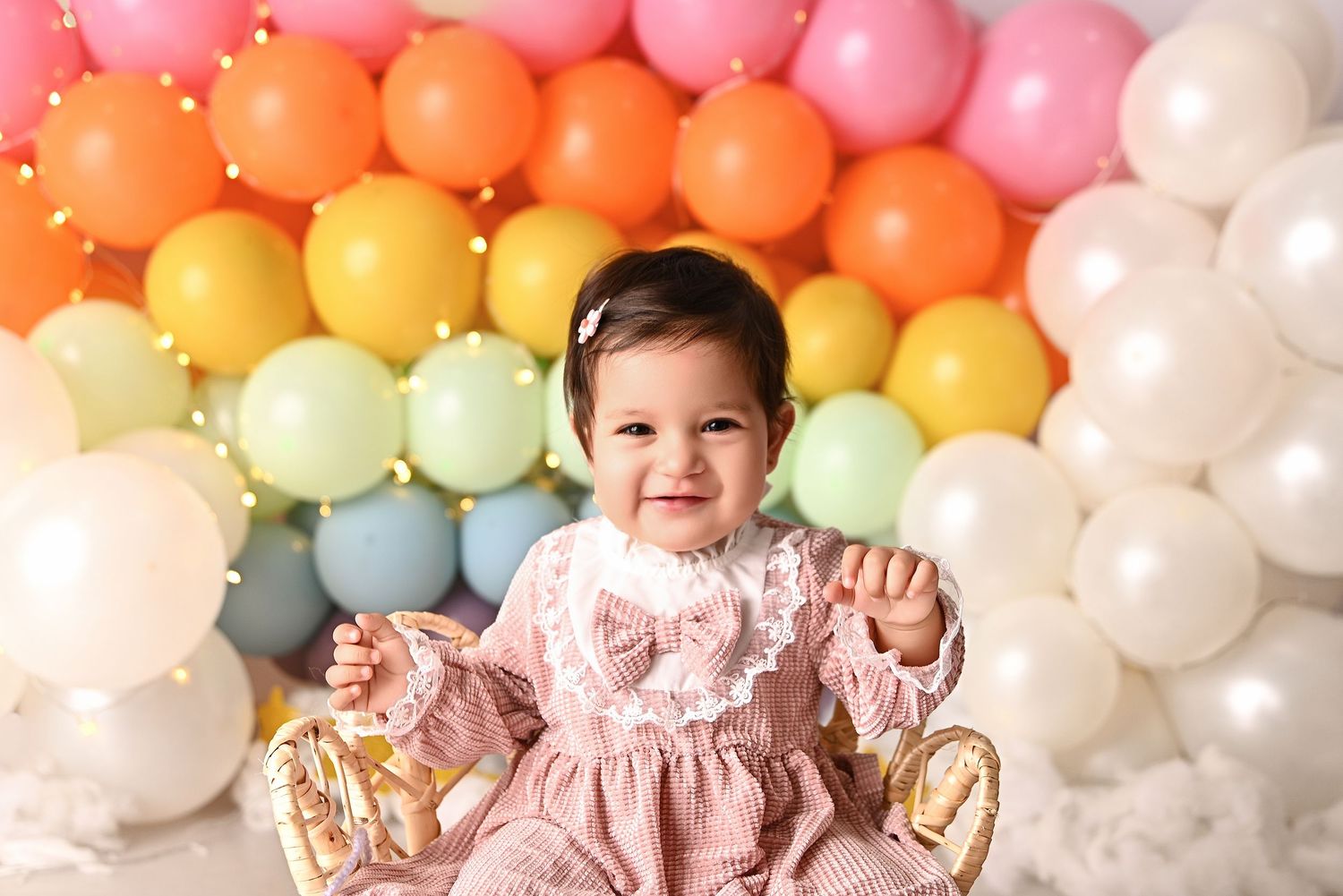 Smiling baby girl in pink romper celebrates first birthday with pastel balloons and number one decoration.