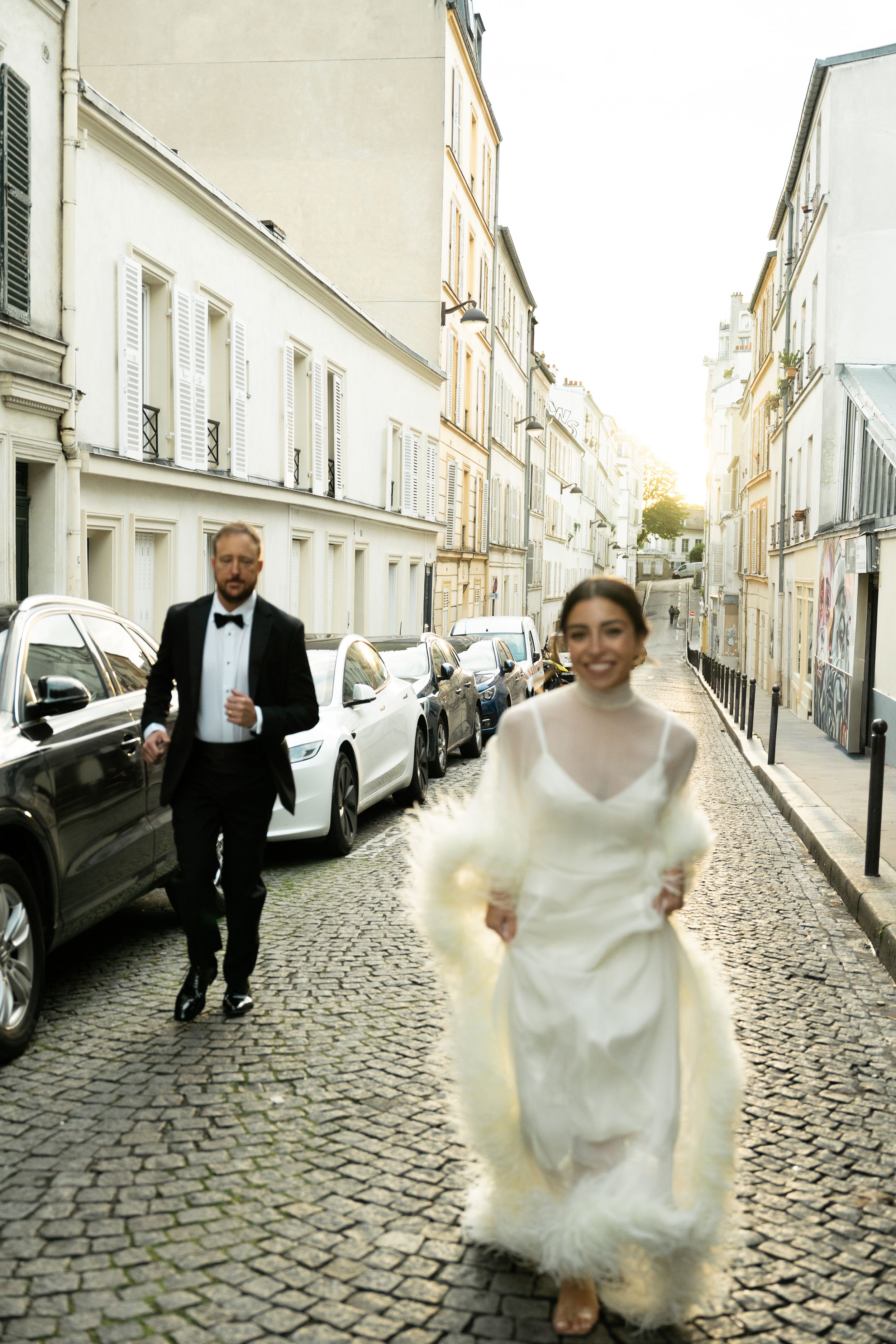 Bride in white gown and groom in tuxedo running on cobblestone street in Paris, France.