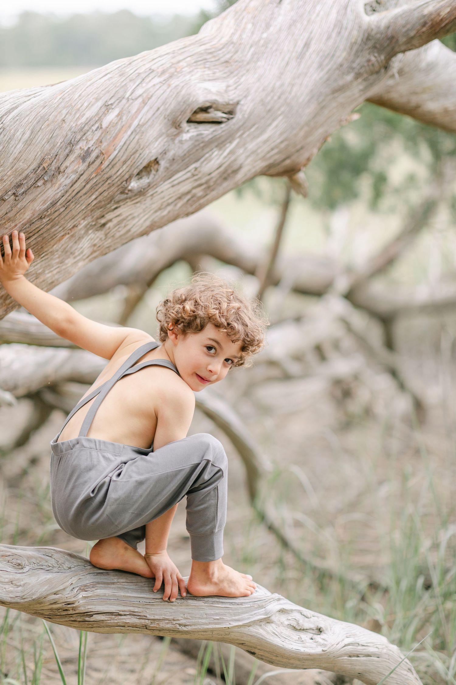 A curly-haired child in gray overalls sits barefoot on a weathered driftwood log in a natural outdoor setting.