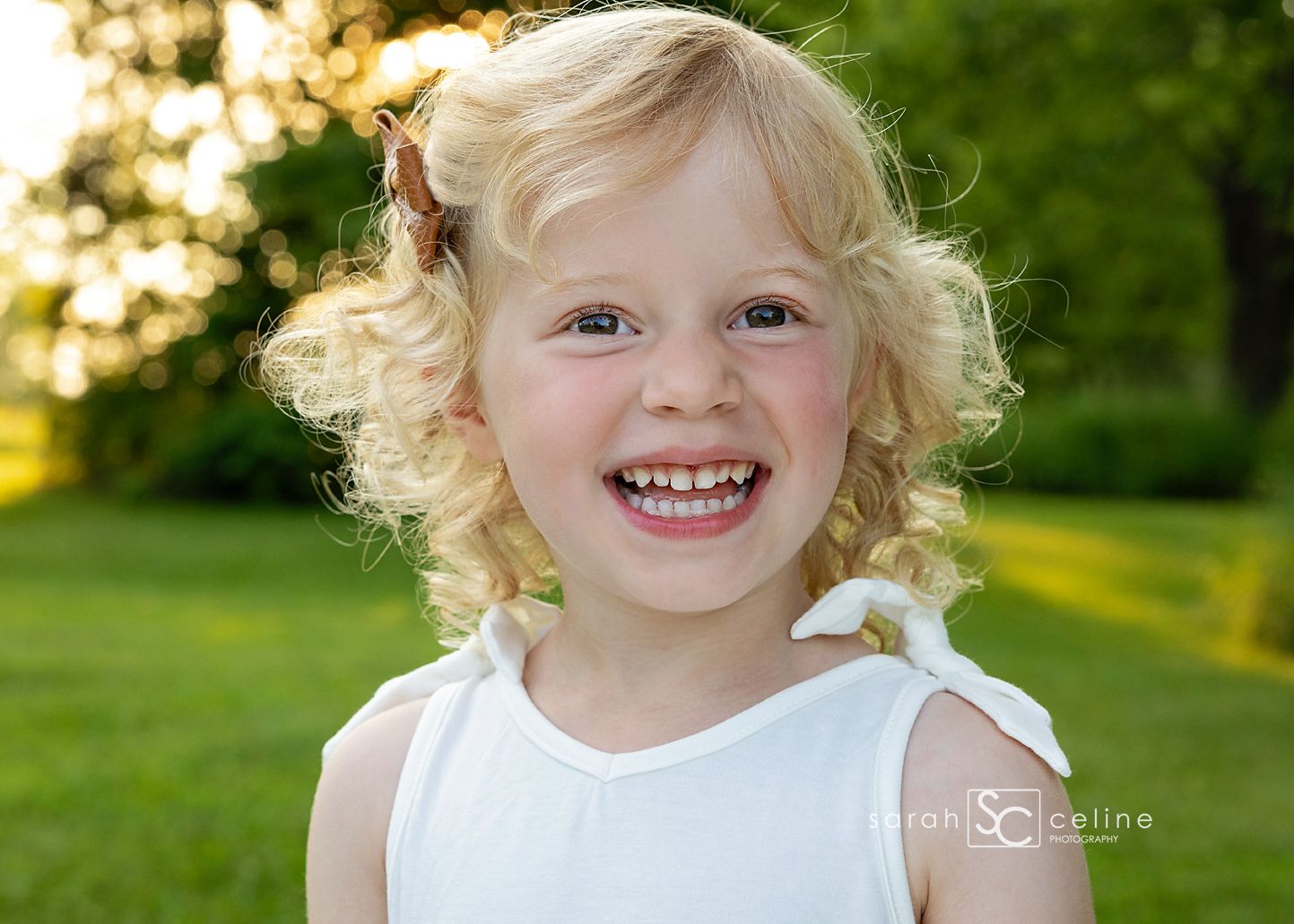 Smiling blonde curly-haired little girl in white dress outdoors in green park with golden sunlight.