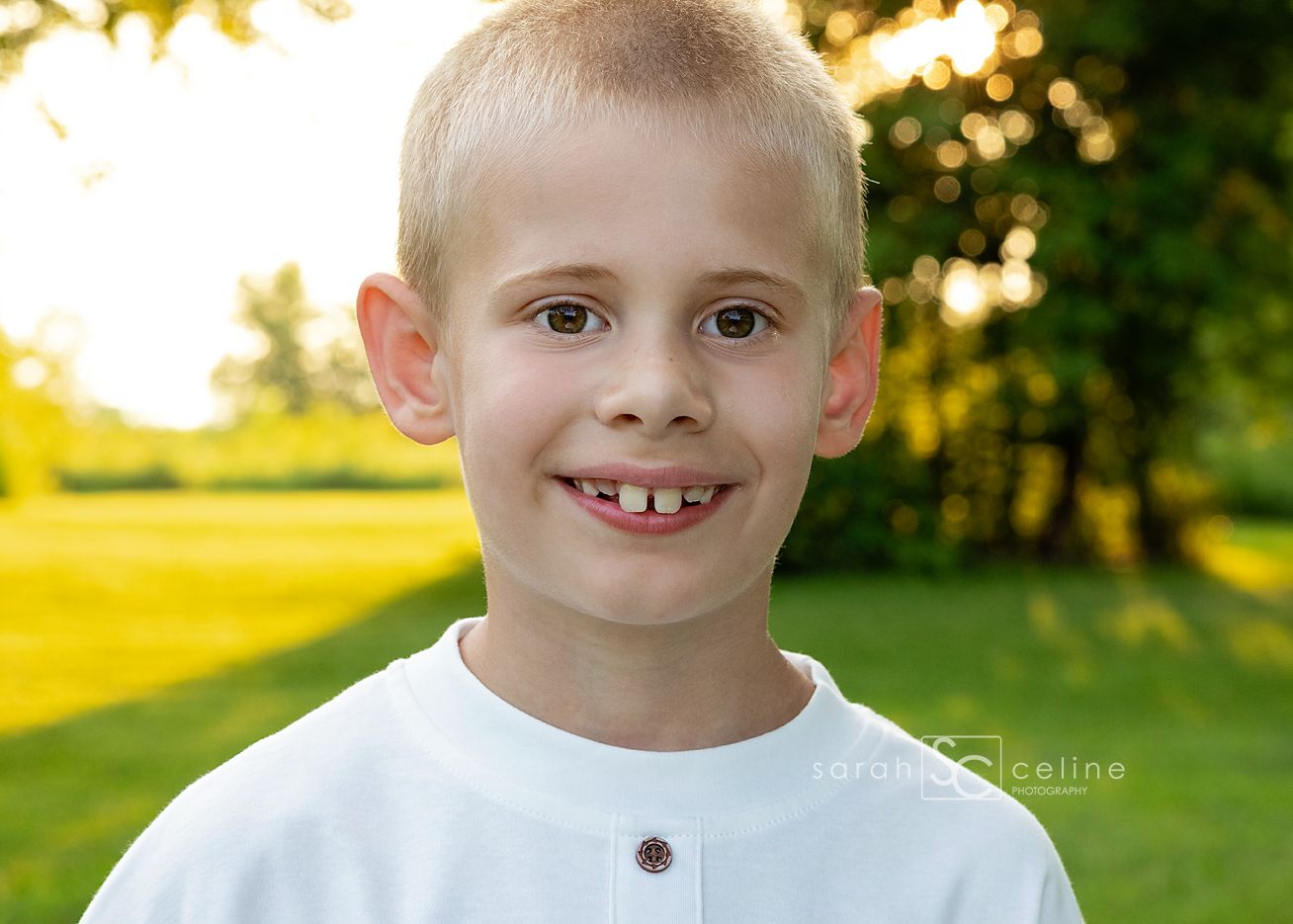 Smiling young boy with blonde hair wearing a white shirt outdoors in a sunny green field.