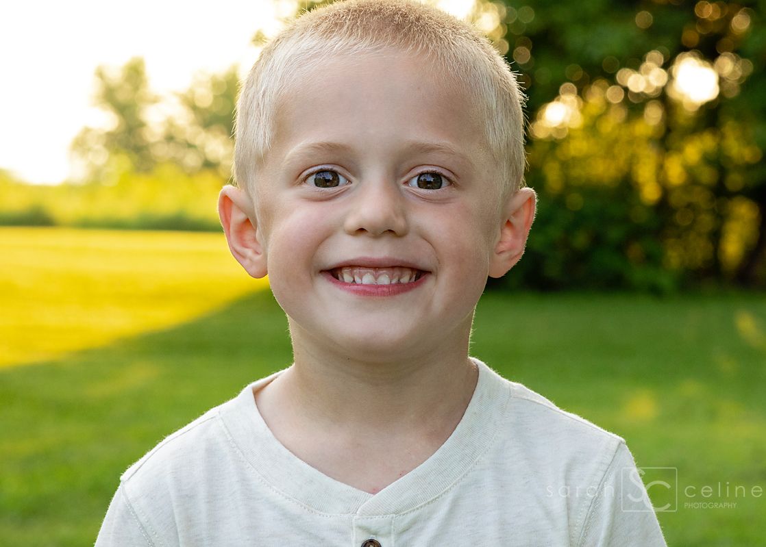 Smiling young blonde boy in a white shirt poses outdoors with green grass and trees in the background.