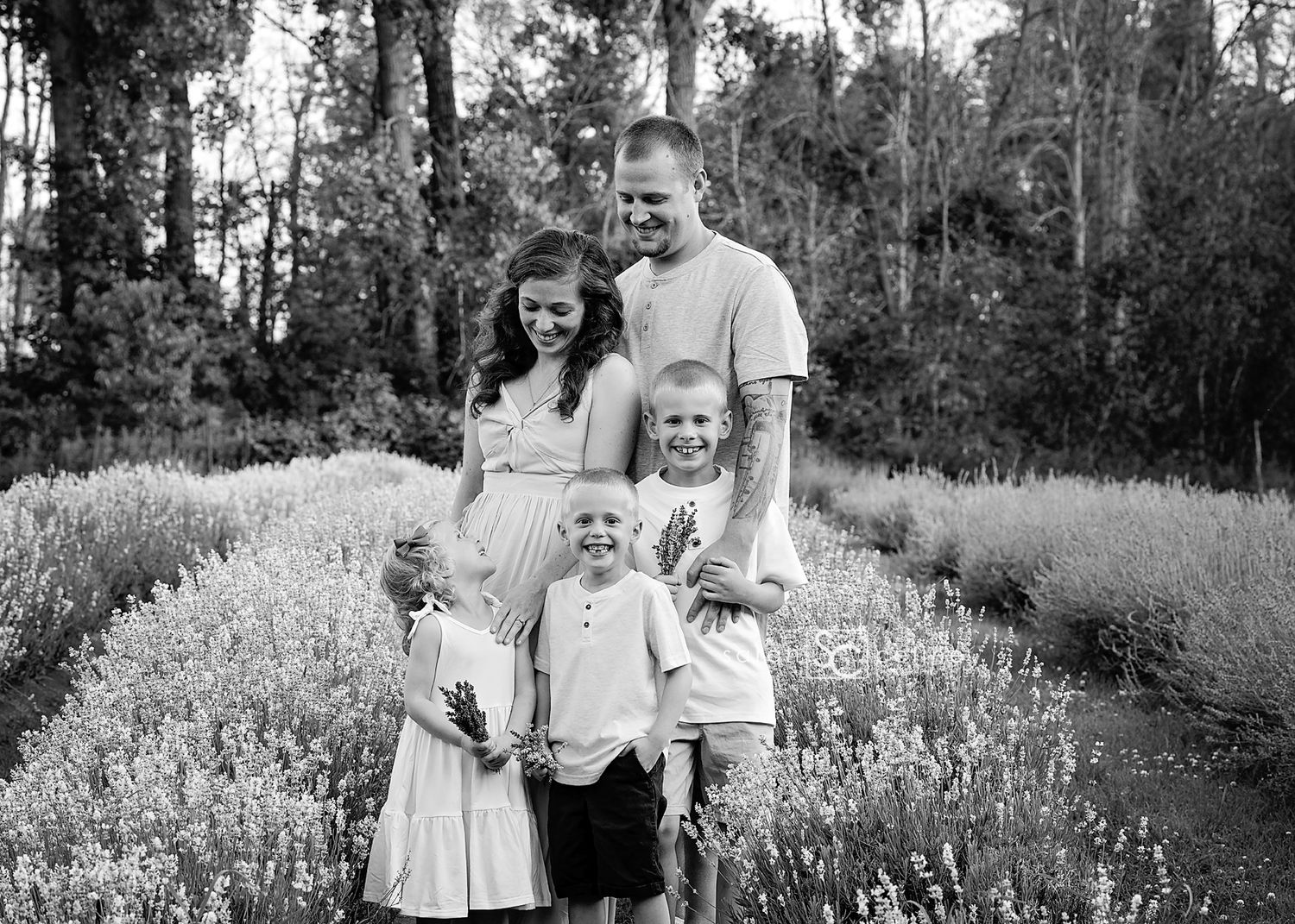 Black and white family portrait of parents with three young children posing in a blooming flower field outdoors.