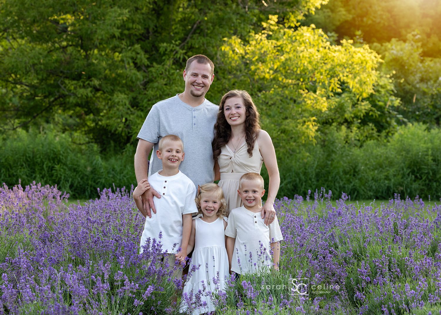 Family of five posing in a purple lavender field with lush green trees in golden hour sunlight.