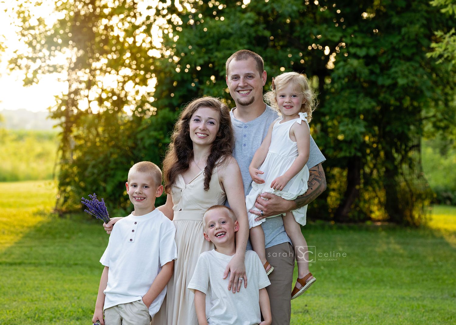 Happy family of five posing outdoors in golden hour sunlight, parents with three young children on lush green lawn.