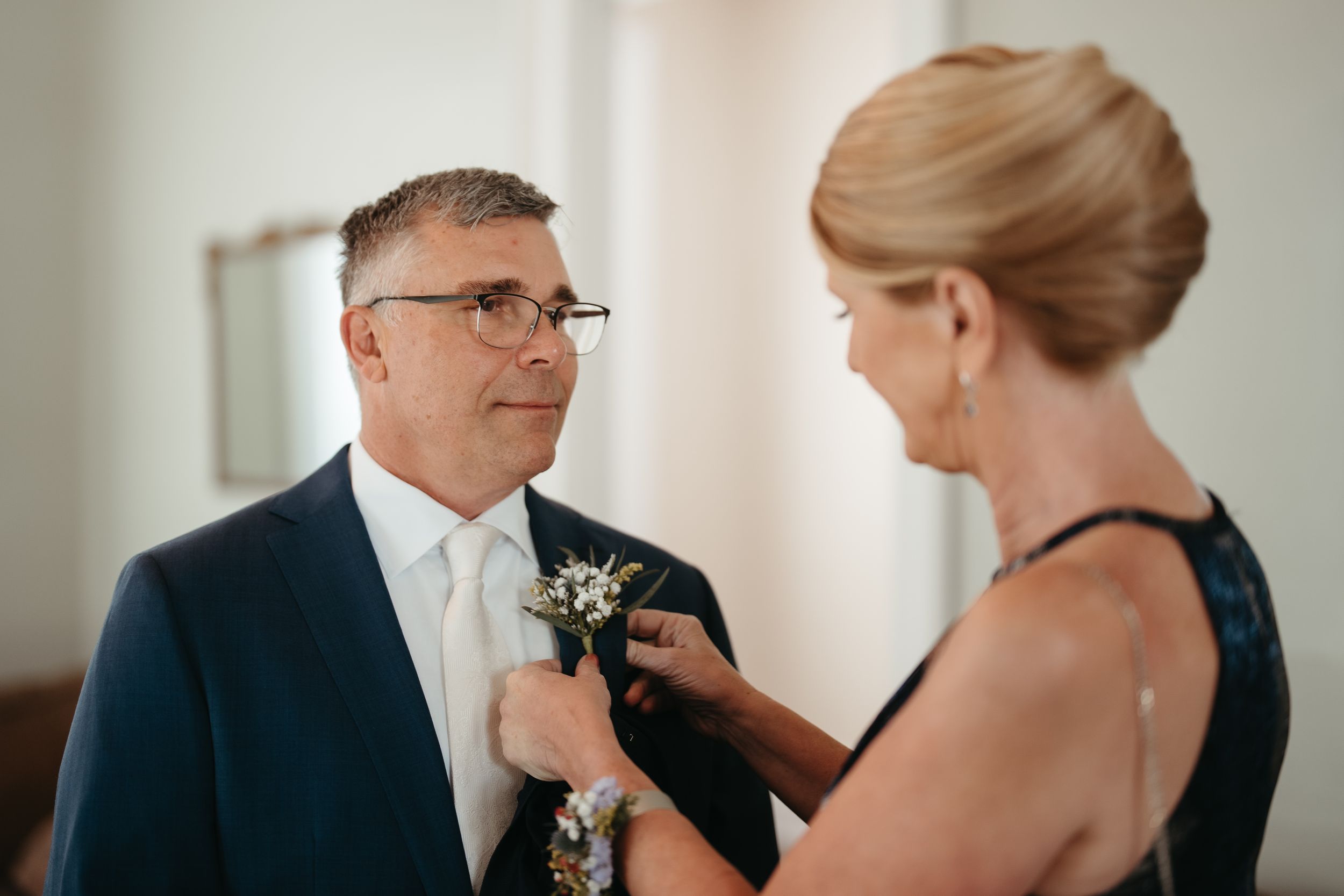 Woman pinning a boutonniere on a man in a navy suit at a wedding preparation.