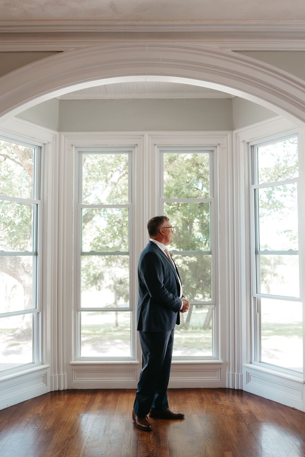 Groom in navy suit stands by large arched windows with hardwood floors in elegant venue.