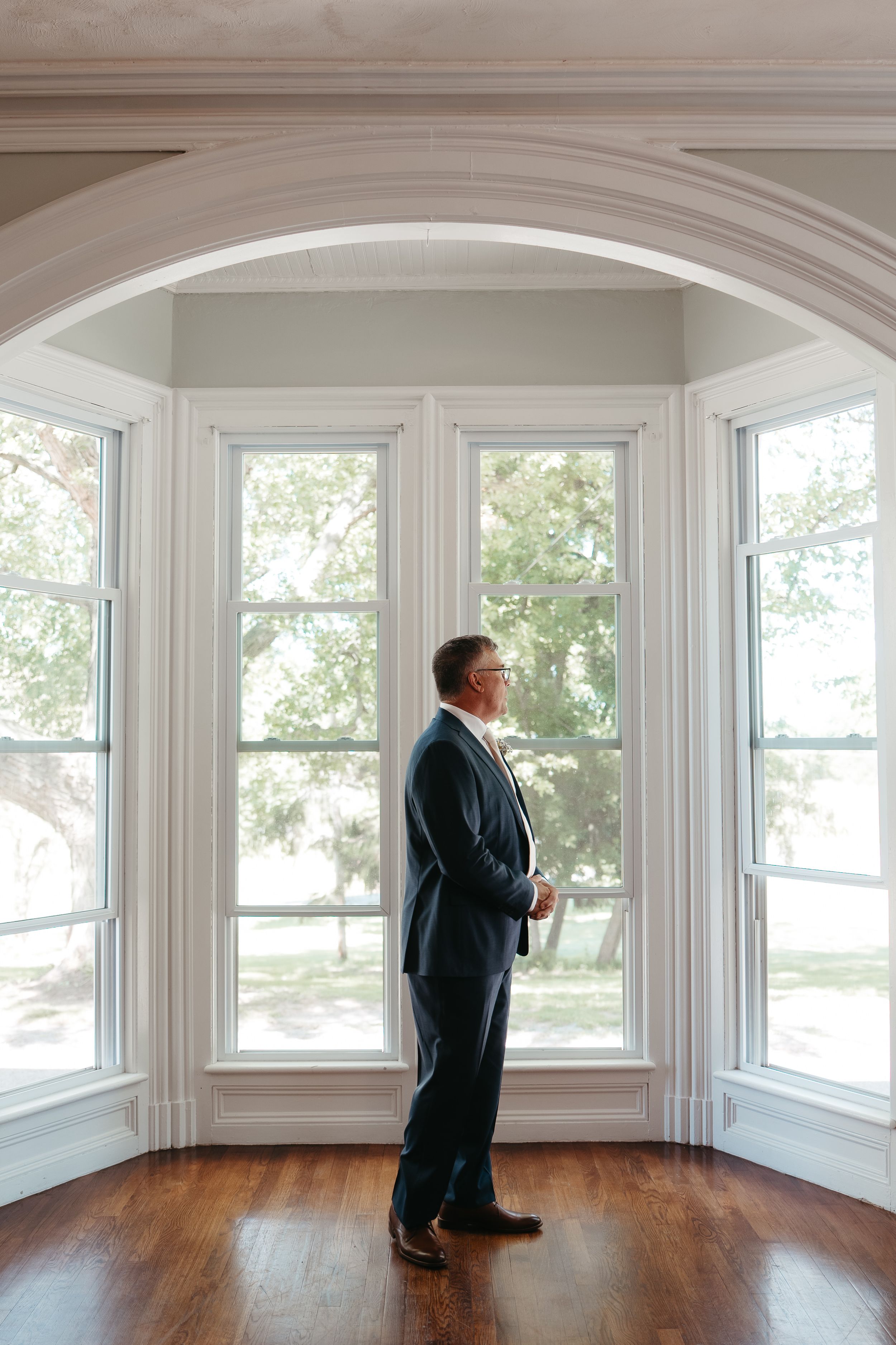 Groom in navy suit stands by large arched windows with hardwood floors in elegant venue.