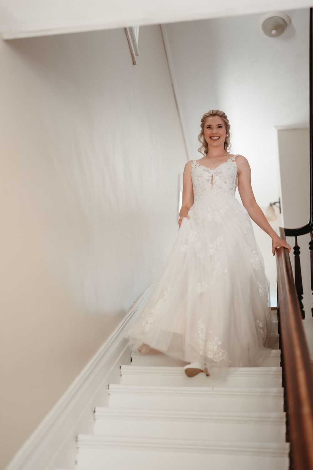 Smiling bride in lace wedding gown descending white staircase with wooden railing.
