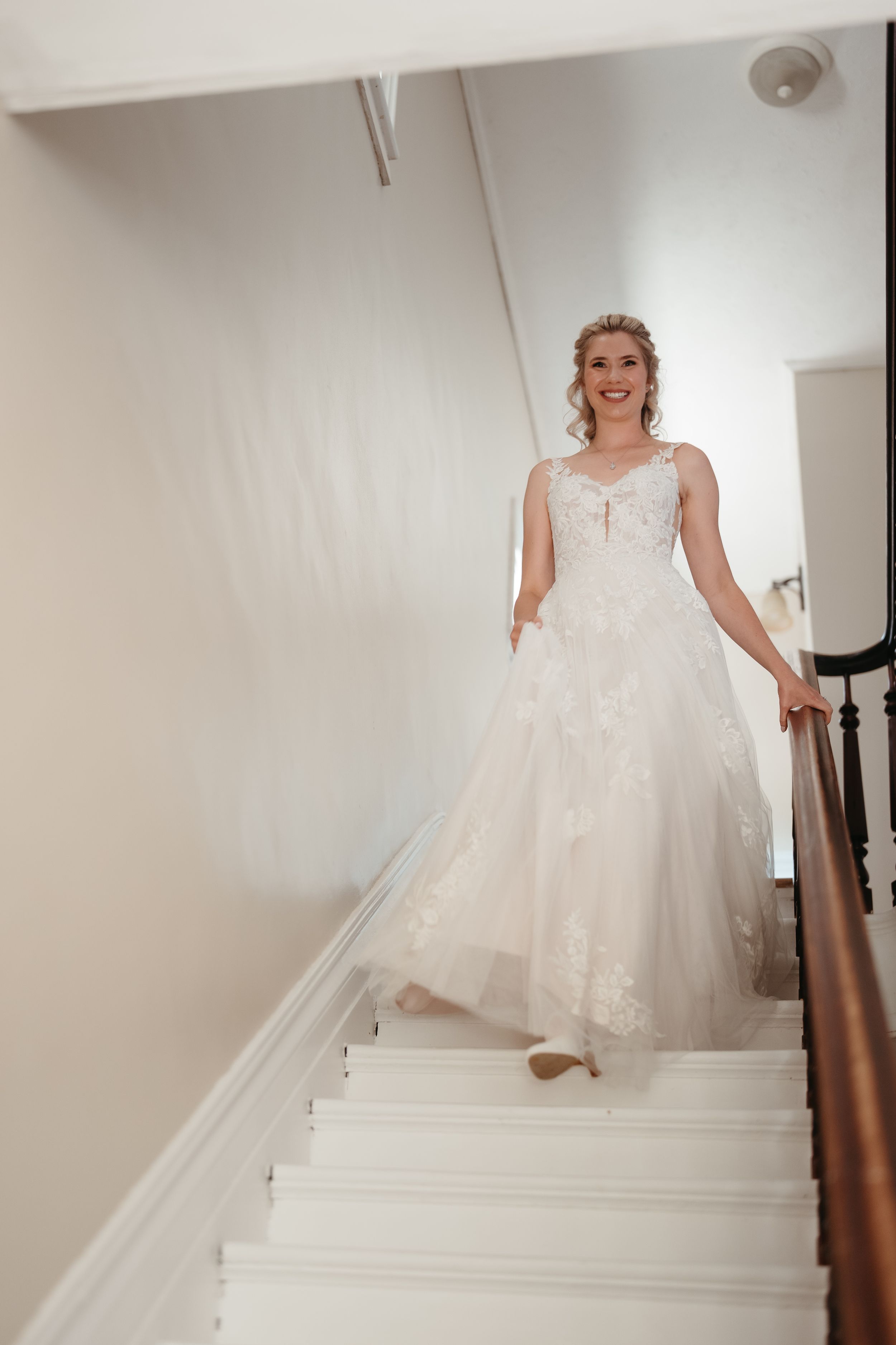 Smiling bride in lace wedding gown descending white staircase with wooden railing.
