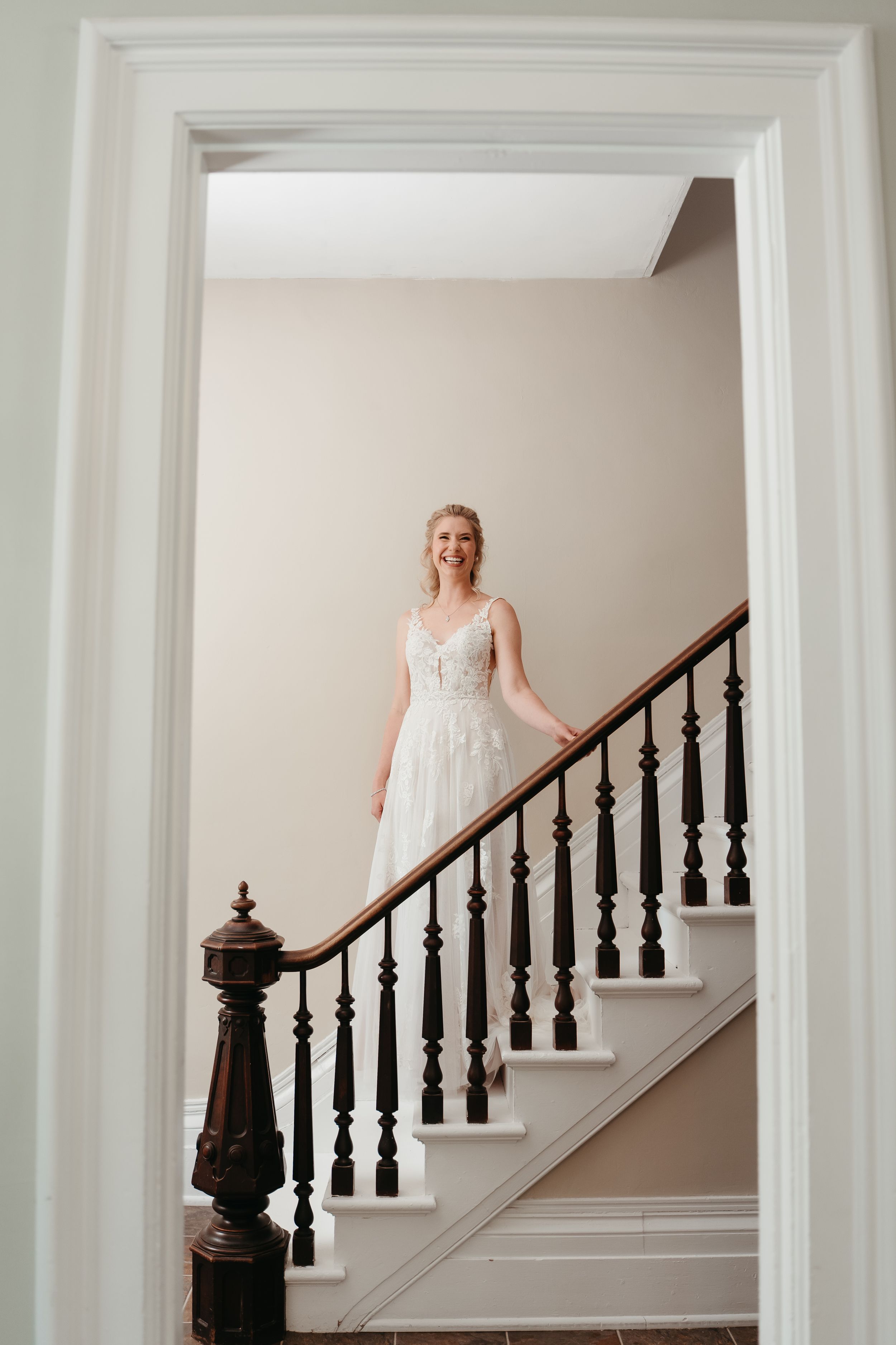 Elegant bride in white lace gown standing on classic staircase framed by doorway.