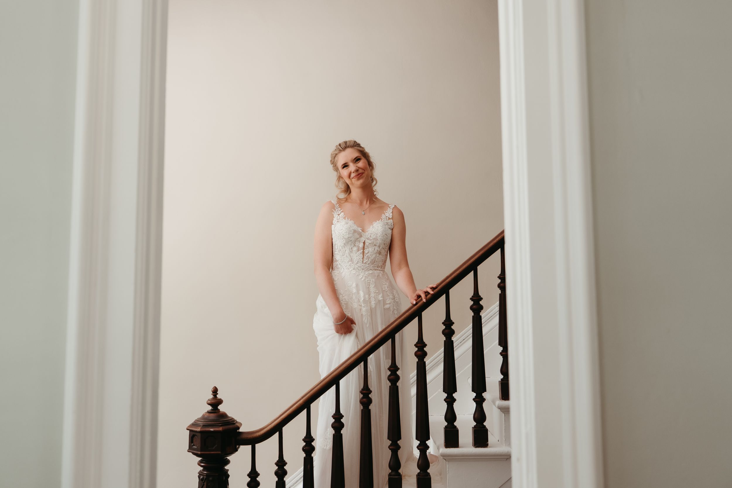 Bride in lace wedding gown smiling on elegant staircase with dark wood railing.