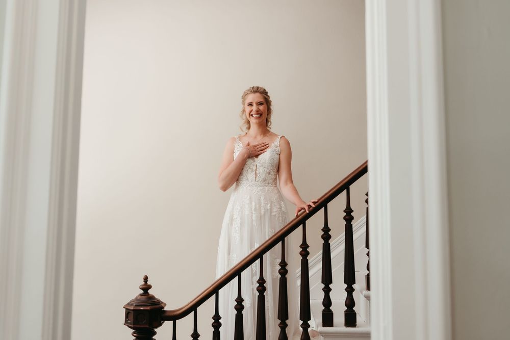 Bride in white lace gown standing on elegant staircase with dark wood railing, smiling gracefully.