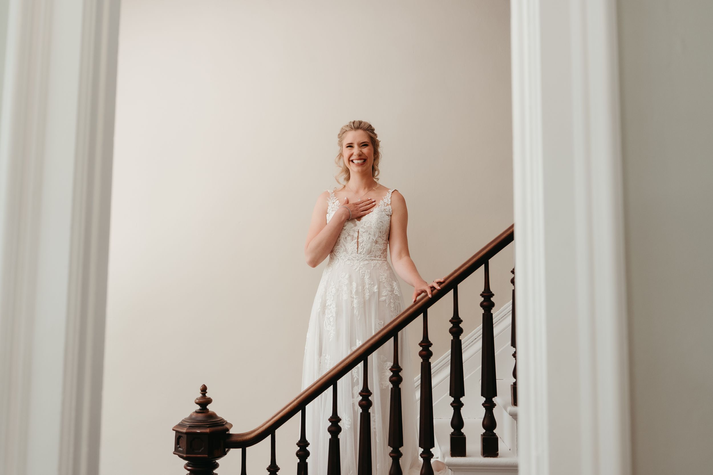 Bride in white lace gown standing on elegant staircase with dark wood railing, smiling gracefully.