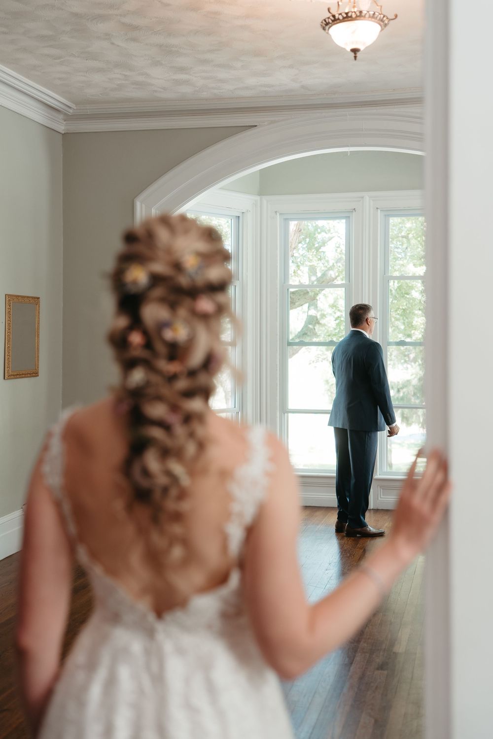 Bride in lace gown with braided updo approaches groom standing at large window in elegant venue.
